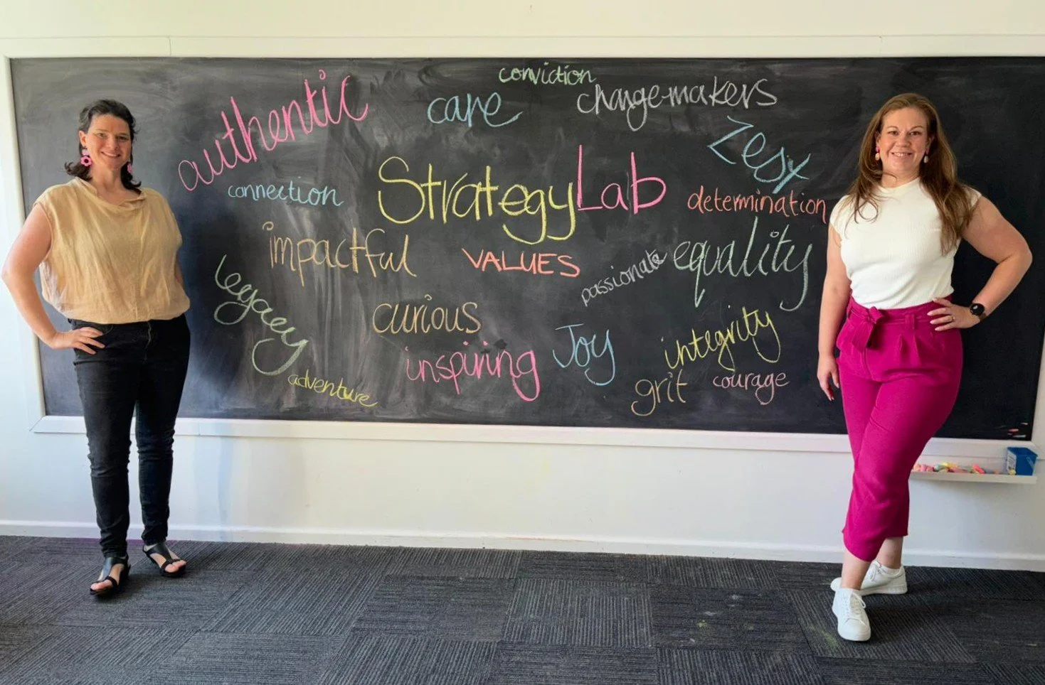 Two women standing on either side of a blackboard titled 'StrategyLab Values', filled with colorful words like 'authentic,' 'impactful,' and 'curious' written in various locations and styles.
