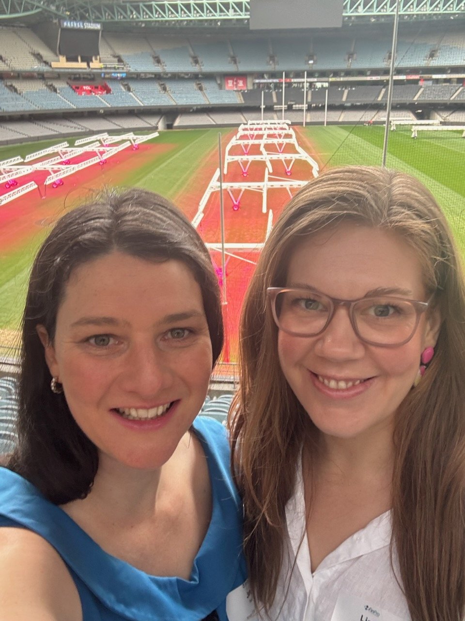 Two women smiling for a selfie inside a stadium's seating area, with a football field and goal posts visible in the background.