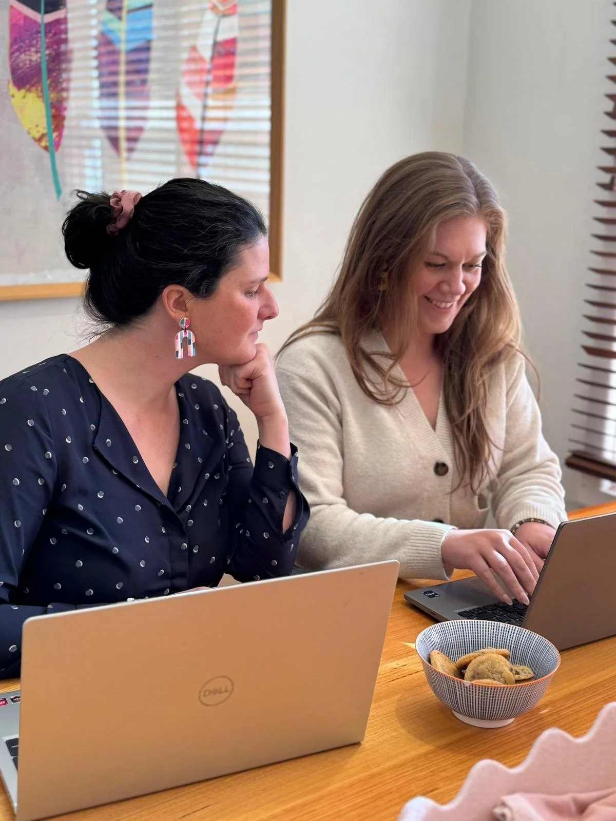 Two women working on laptops at a wooden table with a bowl of cookies, in a bright room with colorful artwork on the wall and blinds.
