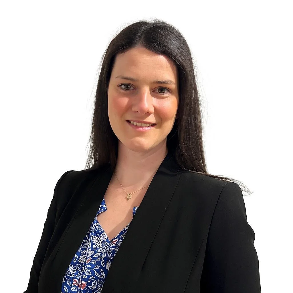 A professional woman with long dark hair, wearing a black blazer and a blue patterned blouse, smiling at the camera against a plain white background.