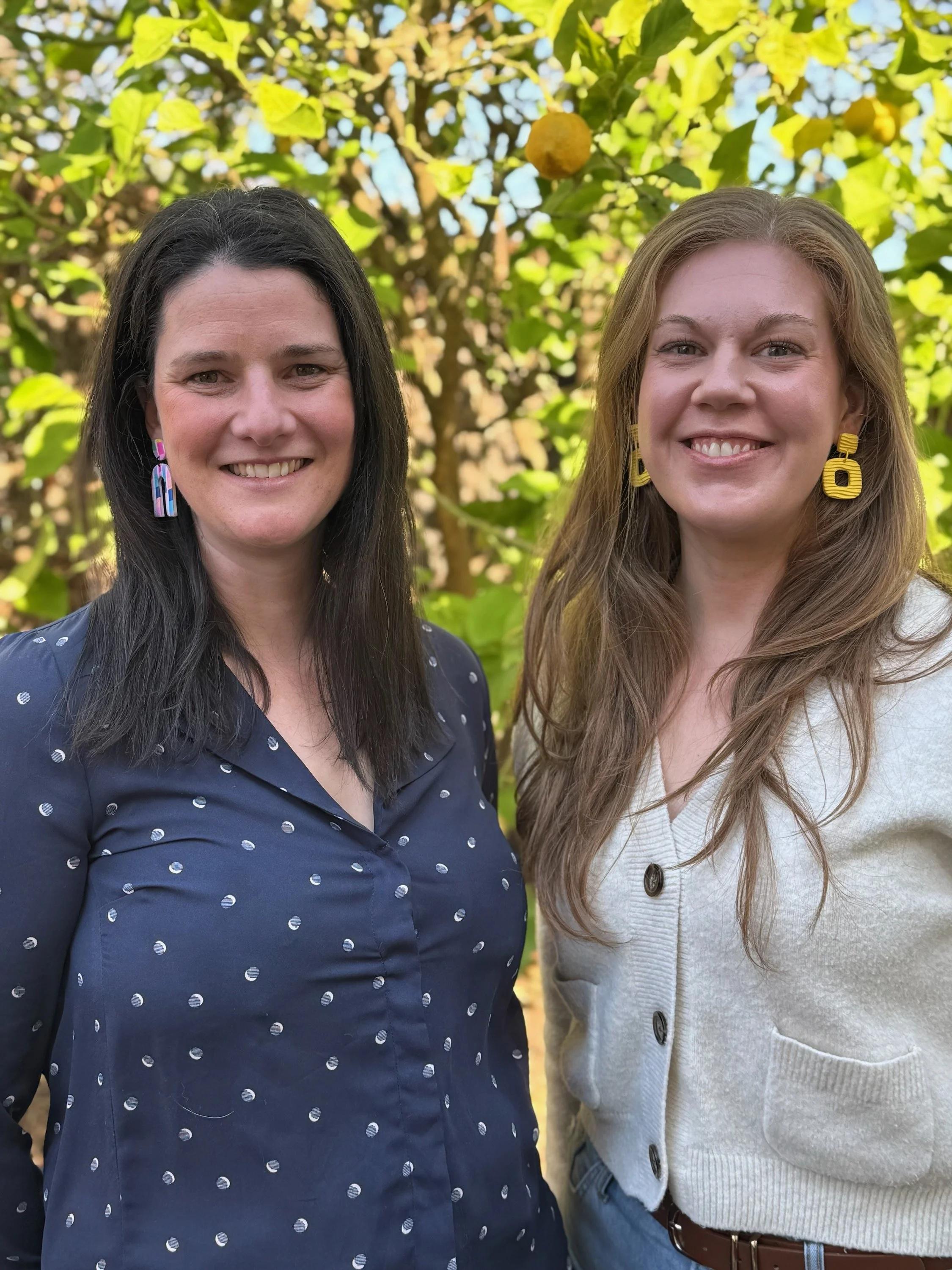 Two women smiling outdoors in front of a tree with yellow and green leaves, one wearing a navy blue polka dot shirt and colorful earrings, the other in a white cardigan with large buttons and yellow earrings.