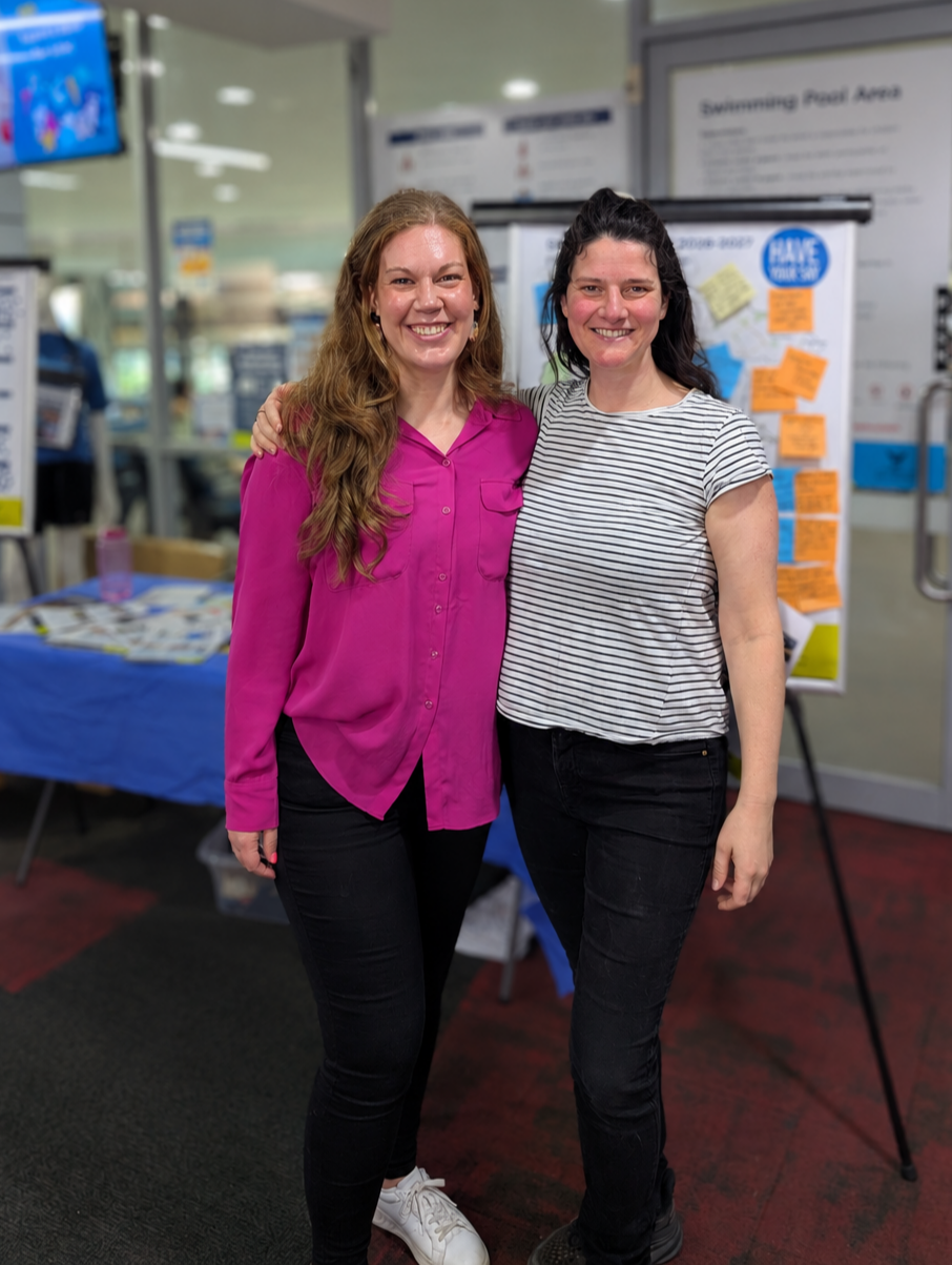 Two women standing close together and smiling, one wearing a pink blouse and the other a striped shirt, in front of a community engagement board with notes, in an indoor setting.