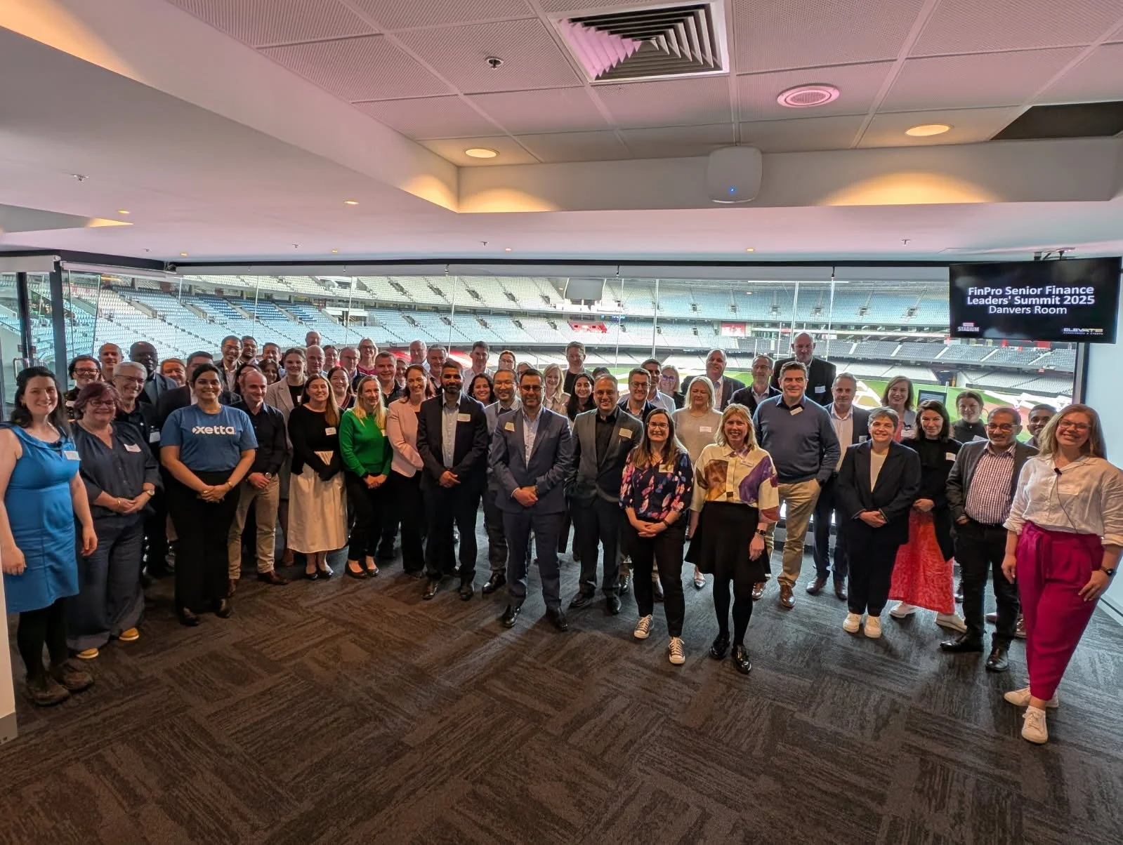 A large group of people, dressed in business attire, gathered in a conference room with a view of a stadium outside the windows. A screen displays information about the event, titled 'FinPro Senior Finance Leaders' Summit 2025 in Danvers Room'.