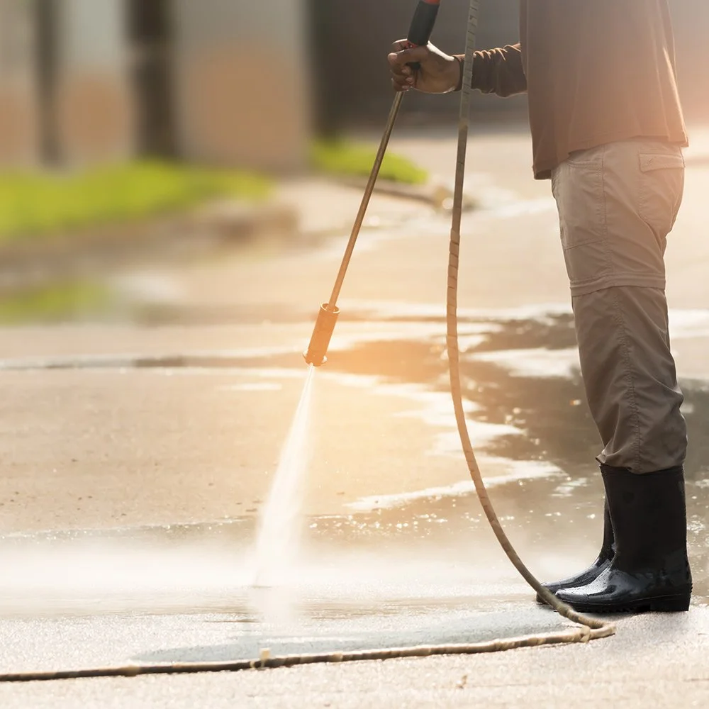 Person pressure washing a concrete sidewalk, wearing black boots and beige pants, with sunlight illuminating the scene.