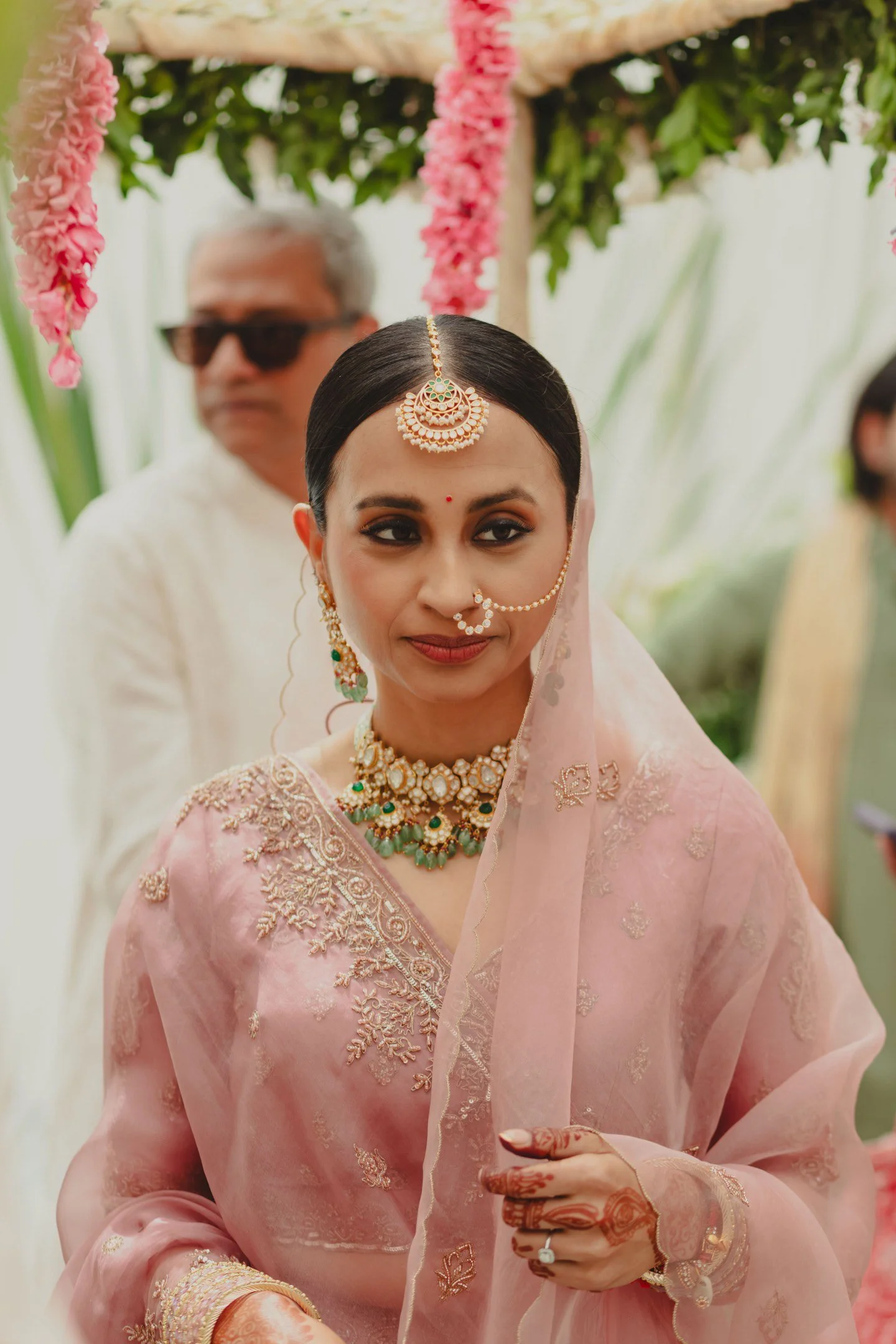 A woman dressed in traditional Indian attire, wearing pink embroidered saree with gold jewelry, including a headpiece, necklace, earrings, and nose ring, standing outdoors during a cultural or wedding ceremony.