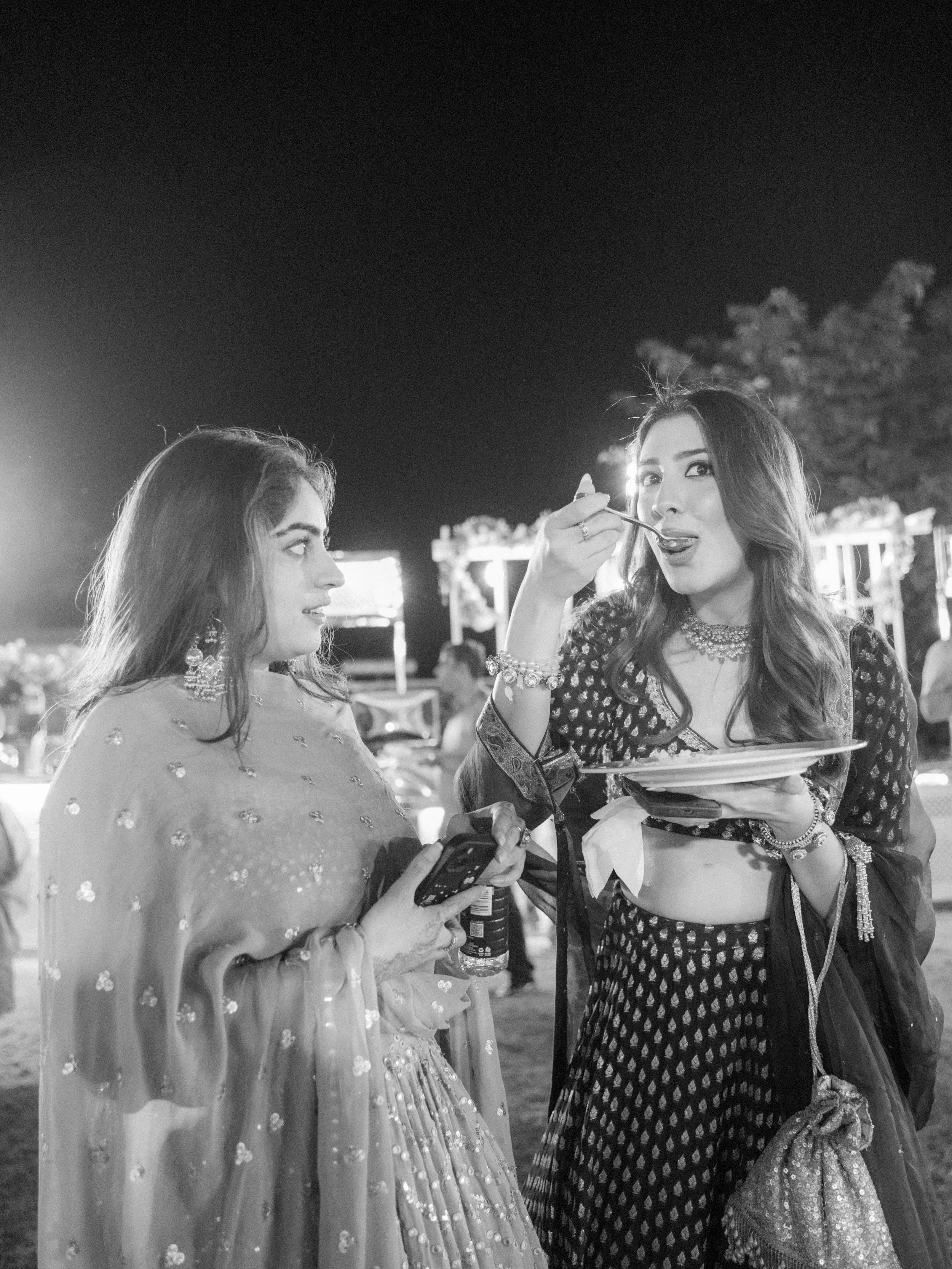 Two women in traditional South Asian attire at an outdoor nighttime event, one eating with a spoon and the other looking at her, with bright lights and trees in the background.