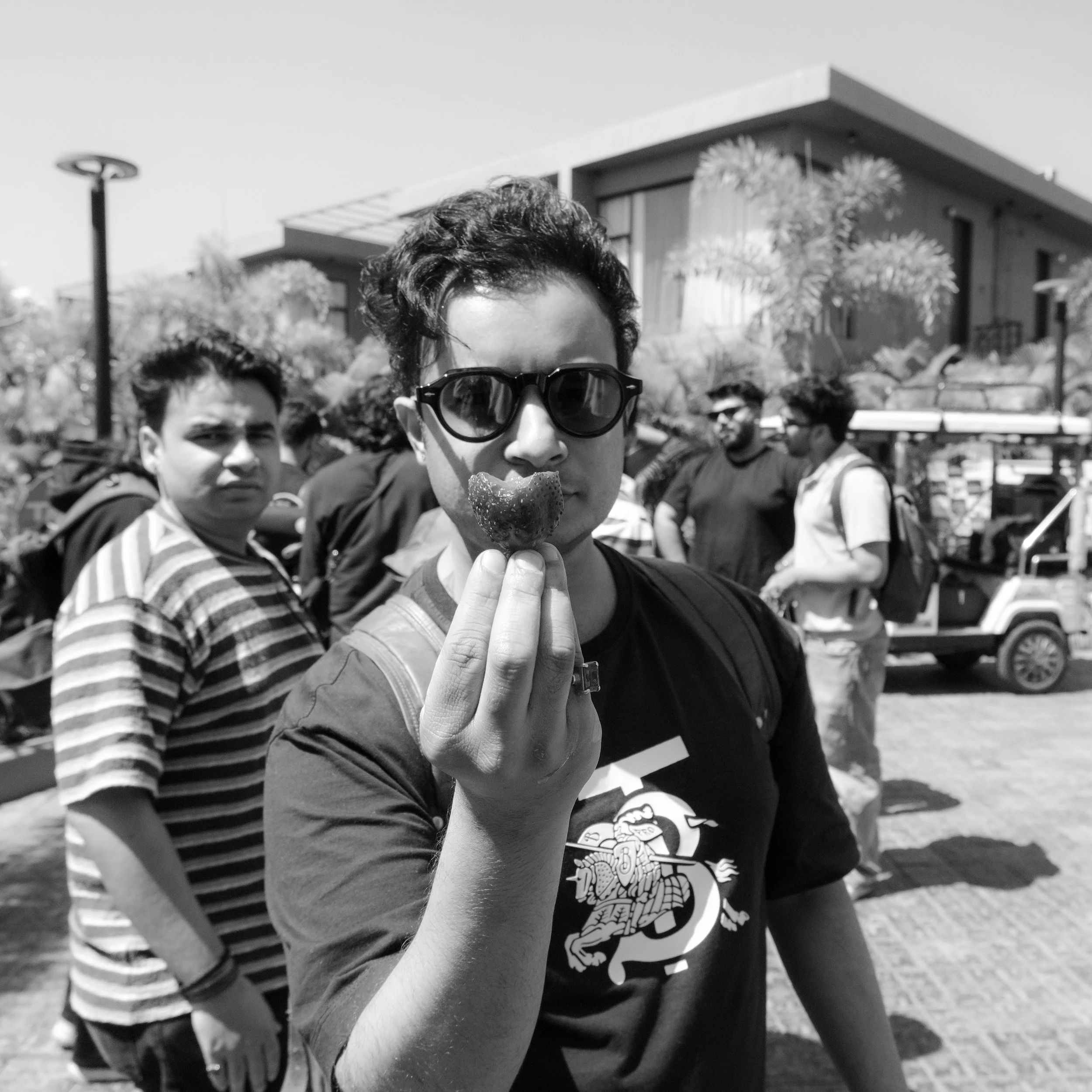 A group of young people outdoors, with a modern house and trees in the background, enjoying a sunny day. The focus is on one person in the front wearing sunglasses and holding a piece of food, while others are visible behind.