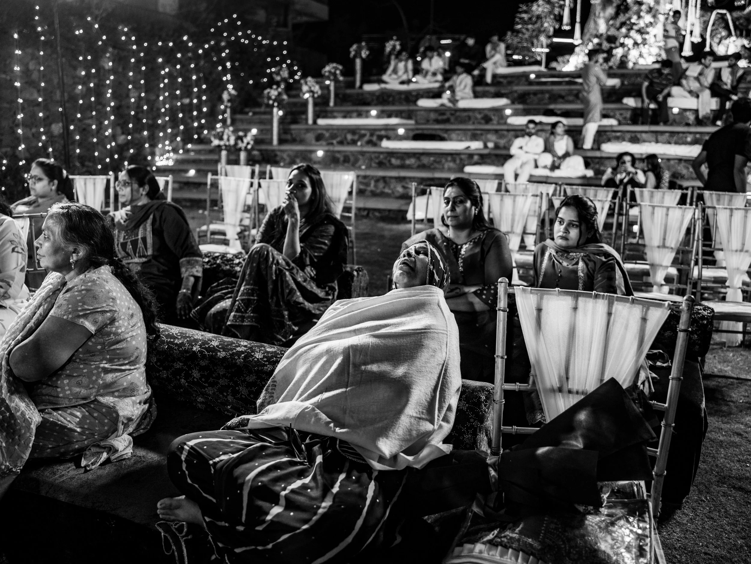 People attending an outdoor night event, some seated on chairs and others on tiered steps, with decorative string lights and flowers in the background.