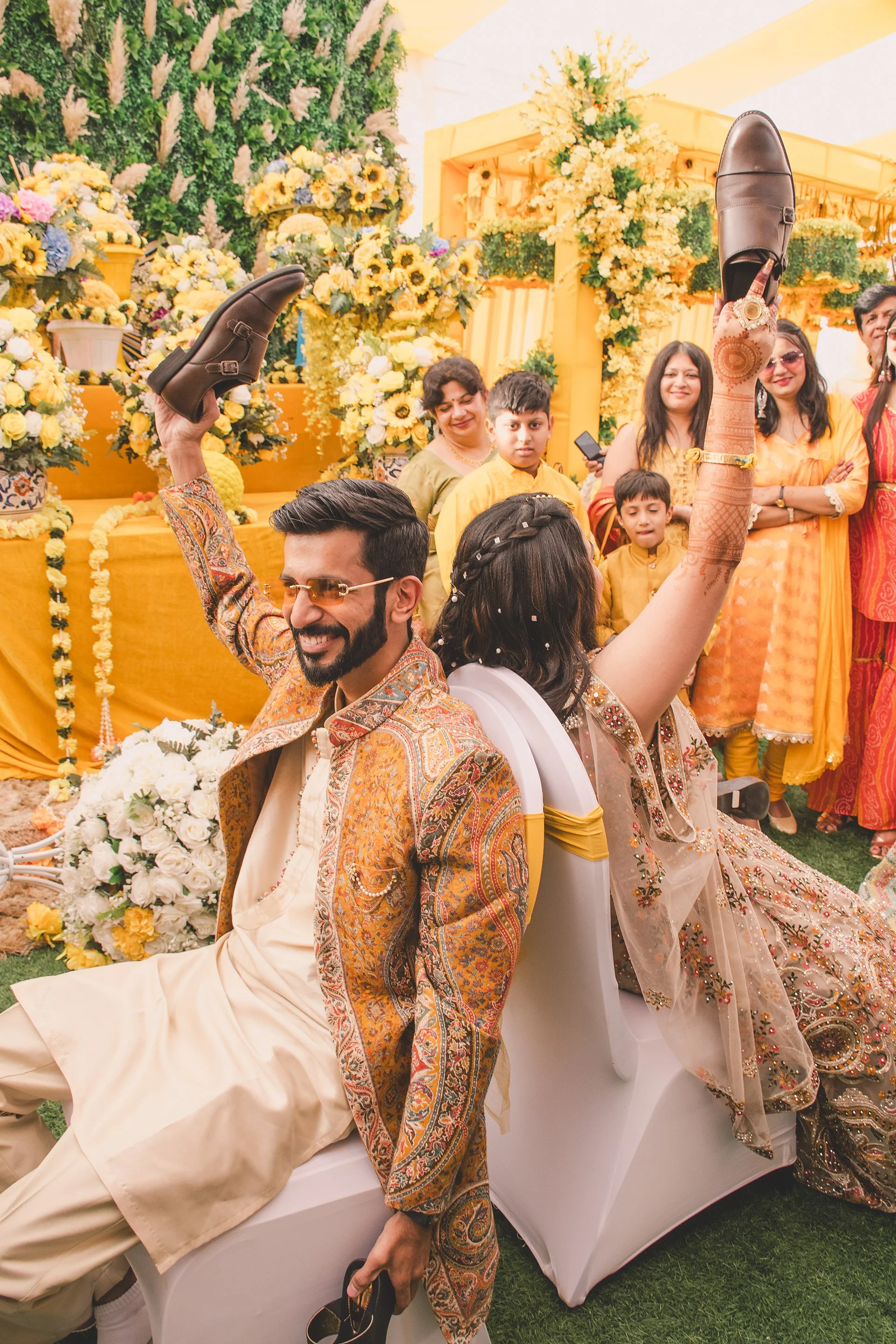 A man and woman seated back-to-back at a celebration, dressed in traditional Indian attire. The woman has her legs raised, showing henna designs, while the man is smiling and holding his shoes. The background is decorated with yellow flowers and peop