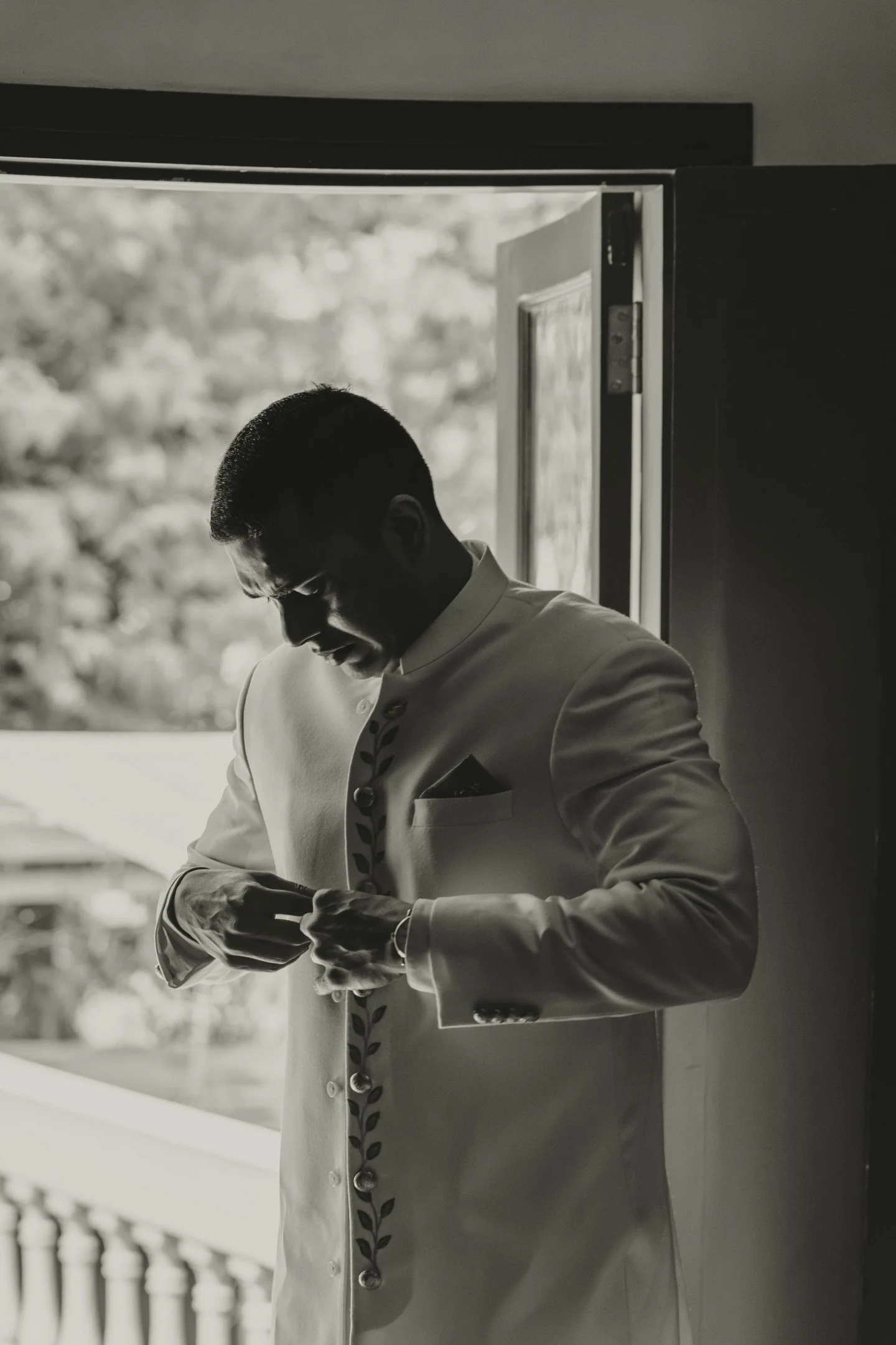 A man wearing a light-colored suit, looking at his phone, standing near a window with an outdoor view of trees, in black and white.