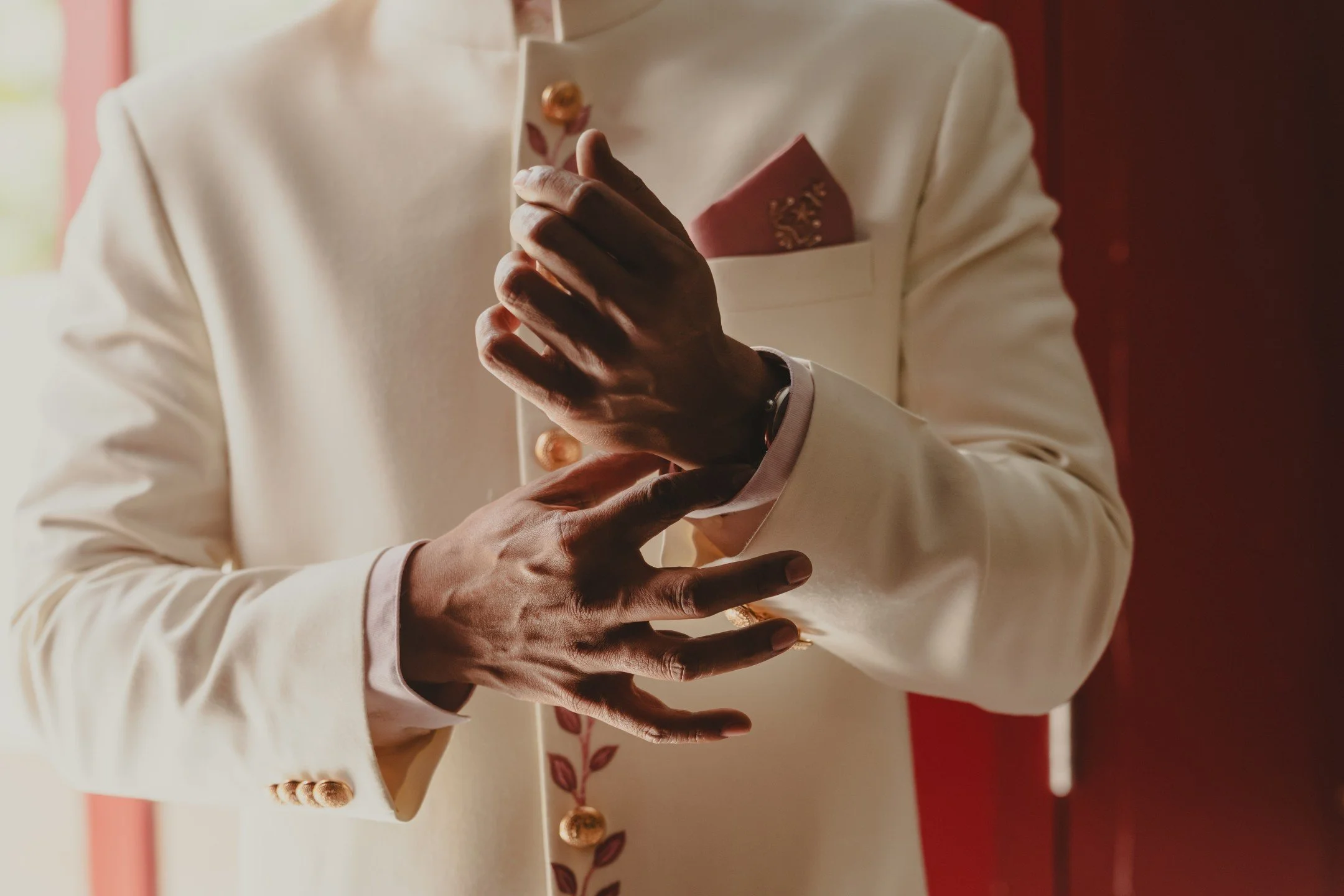 A person dressed in a white suit with gold buttons is holding a smartphone in their hands. The background features red and white colors with floral patterns.