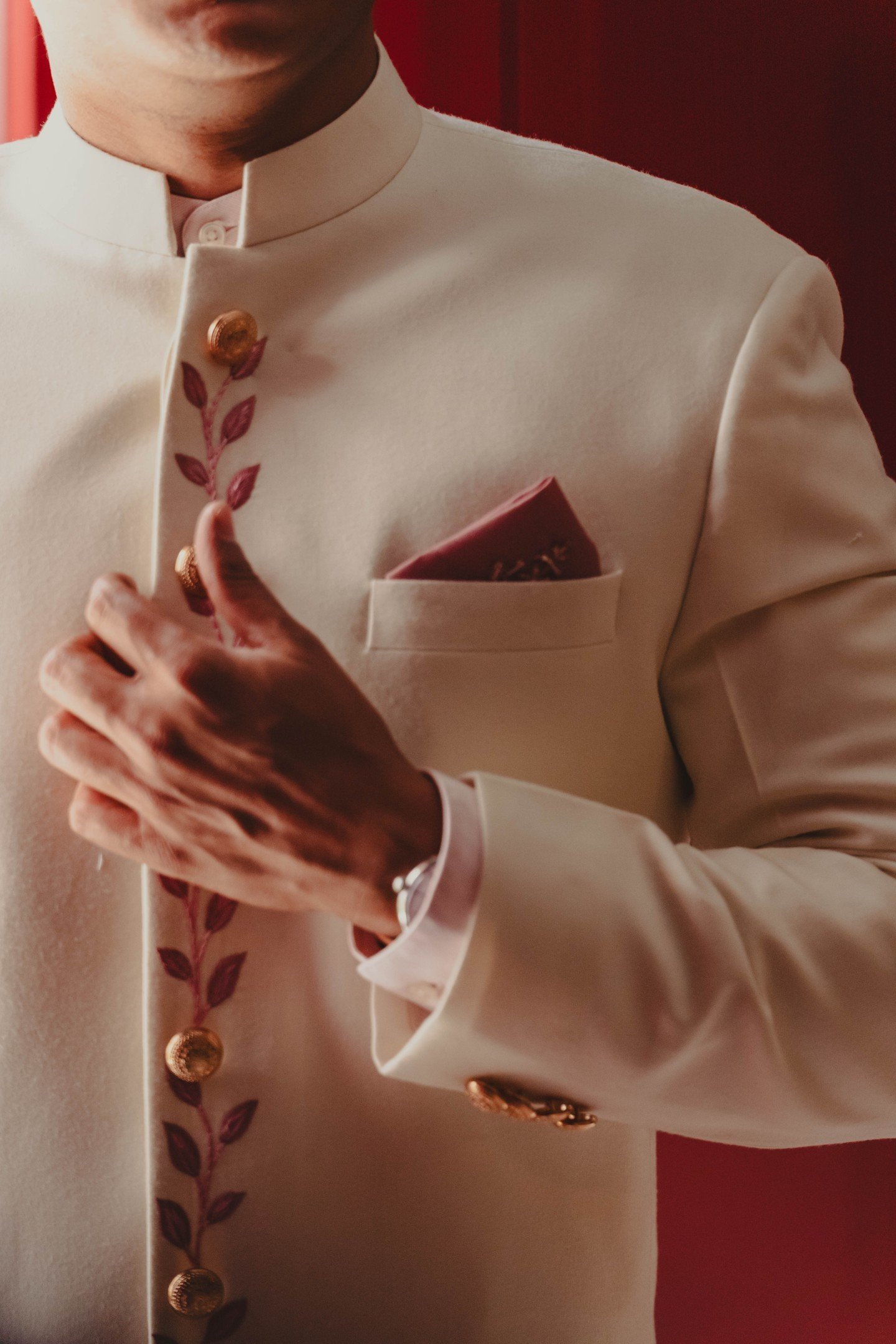 Close-up of a person wearing a cream-colored, traditional South Asian kurta with gold buttons, a decorative pocket square, and a silver wristwatch, set against a red background.