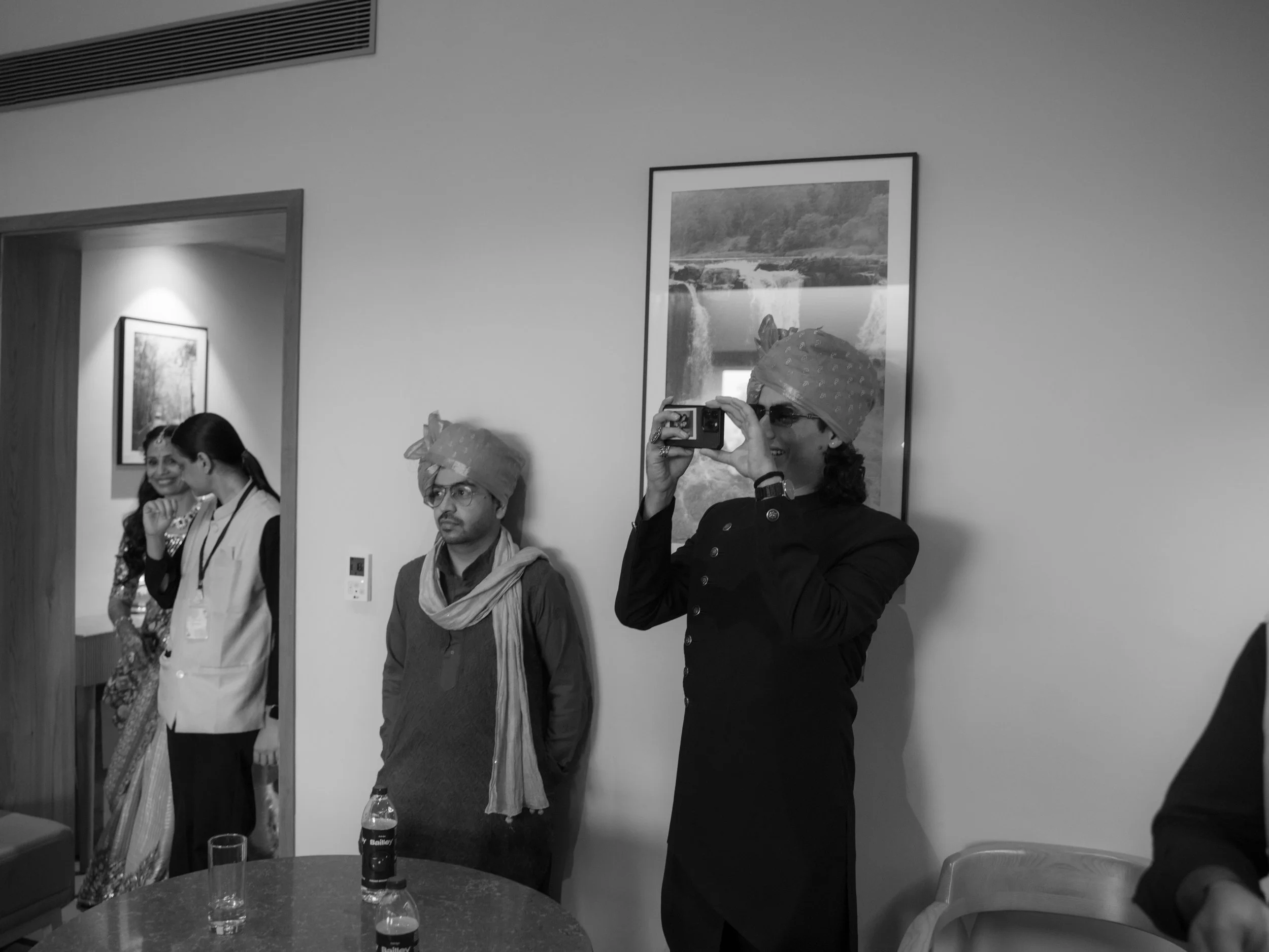 People gathered at a social event, some taking photos and others conversing, with framed pictures on the wall and glasses on a table.