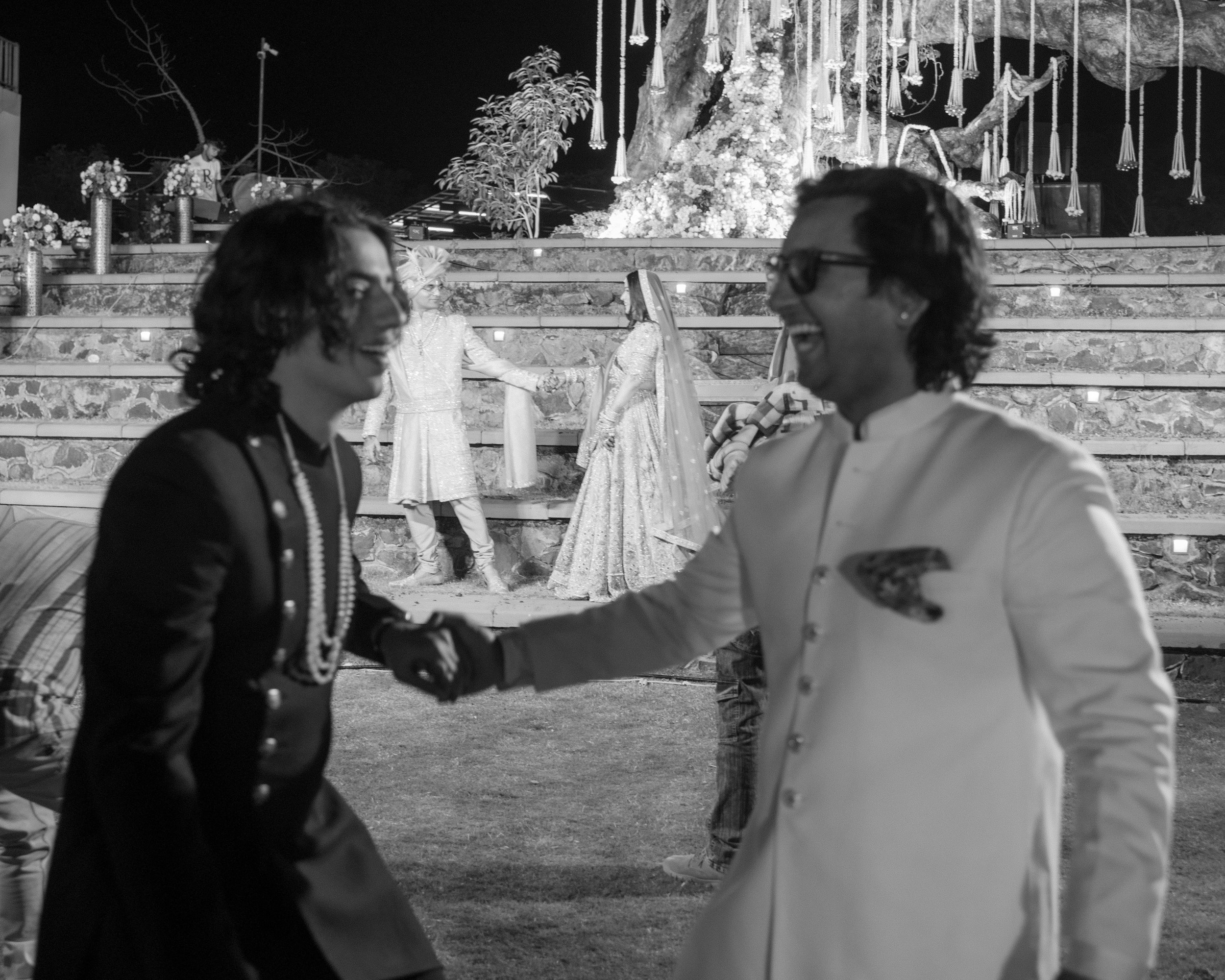 A black and white photo of a wedding ceremony at night. Two people are in the foreground, holding hands and smiling at each other. In the background, there are three women dressed in traditional attire, with one in the center performing the ceremony.
