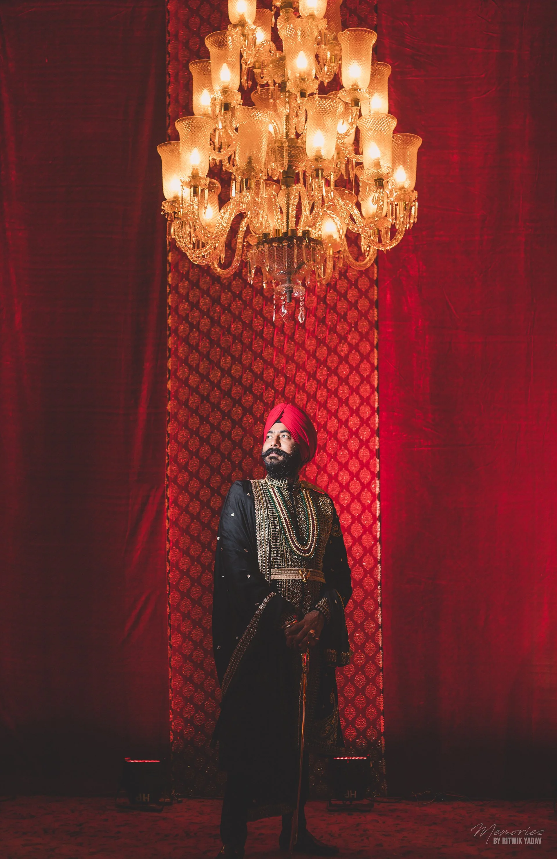 A man dressed in traditional Indian attire, including a red turban, black jacket with intricate designs, and jewelry, stands in front of a red backdrop with patterned curtains. He is holding a staff and looking to his left, beneath a large ornate chandelier with multiple lit bulbs hanging from the ceiling.