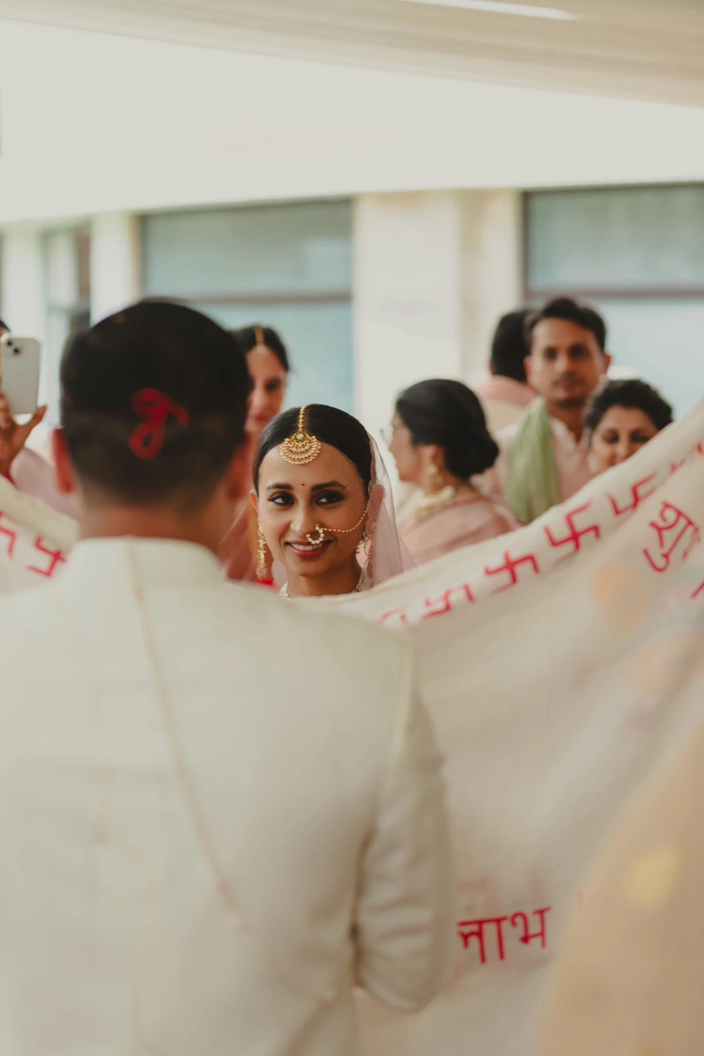 A woman dressed in traditional Indian attire, wearing jewelry including a maang tikka and nose ring, stands amidst a crowd at a cultural or religious event. The crowd features people wearing sarees and kurtas, with some partially visible adults and c