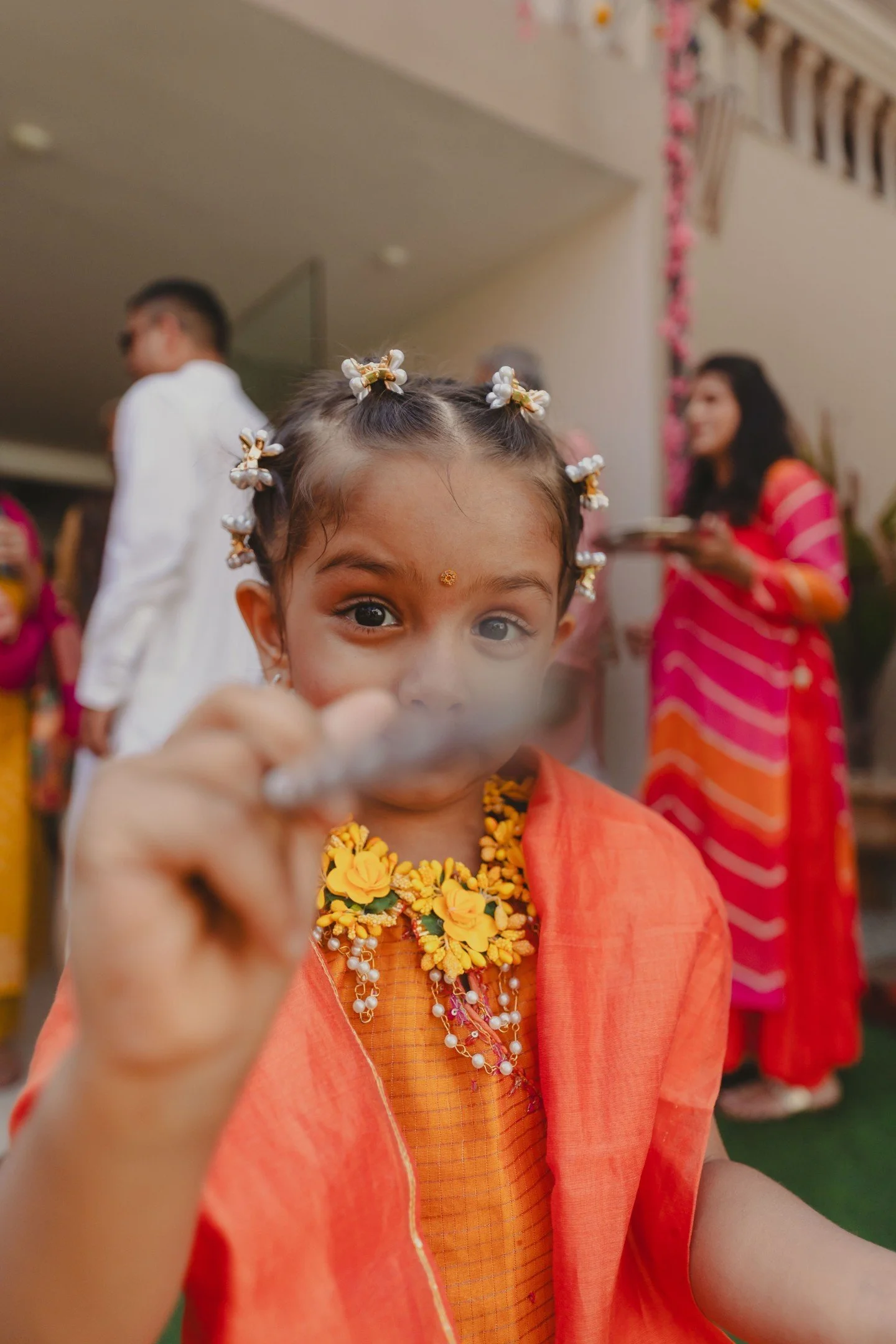 A young girl with braided hair adorned with white and gold beads, wearing orange traditional attire with floral jewelry and a gold decorative bindi on her forehead, participating in a celebration with other people dressed in colorful clothing in the background.
