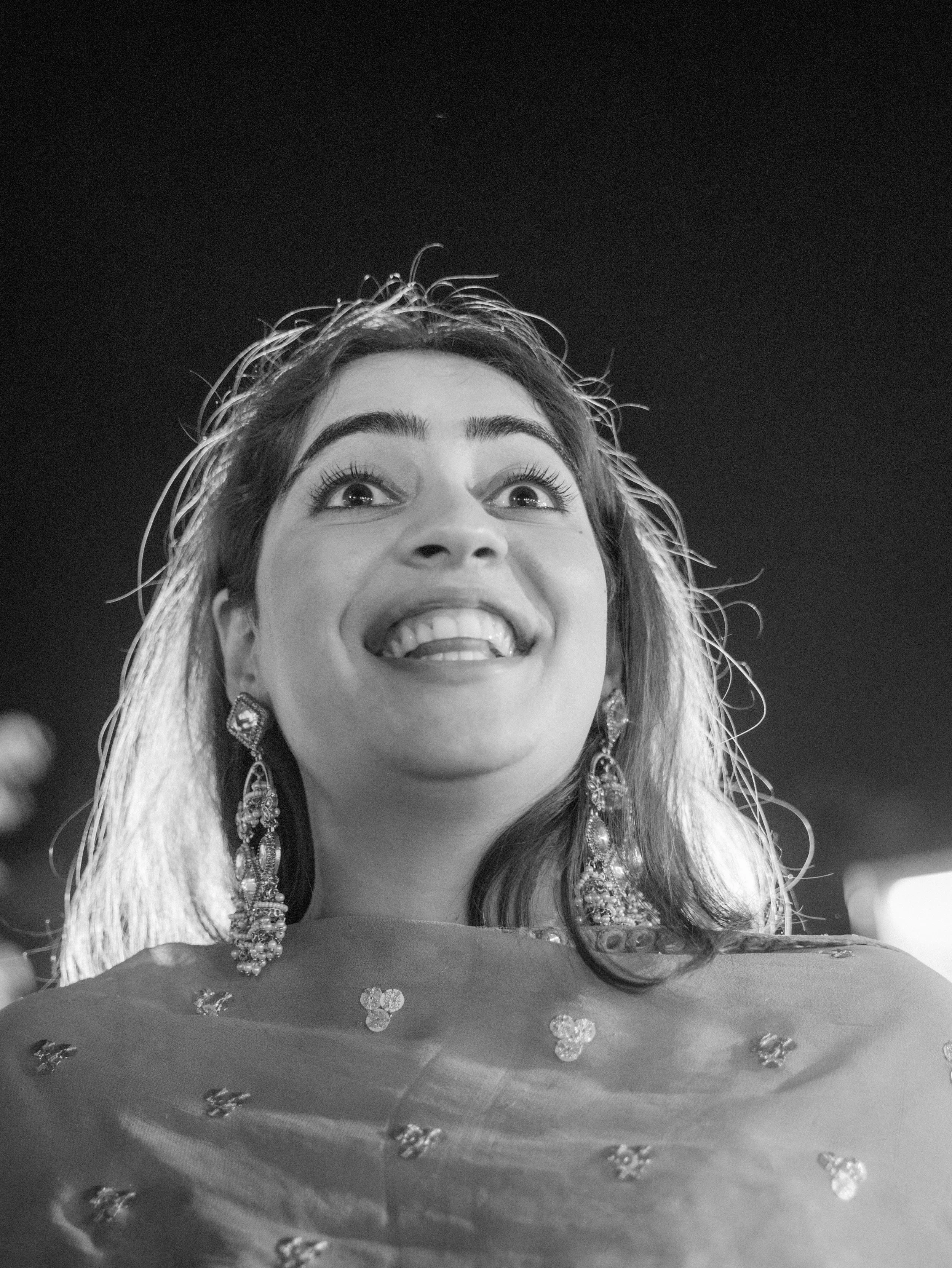 A woman smiling and looking upwards, wearing earrings and a dress with floral embellishments, with her hair styled loosely, against a dark background.