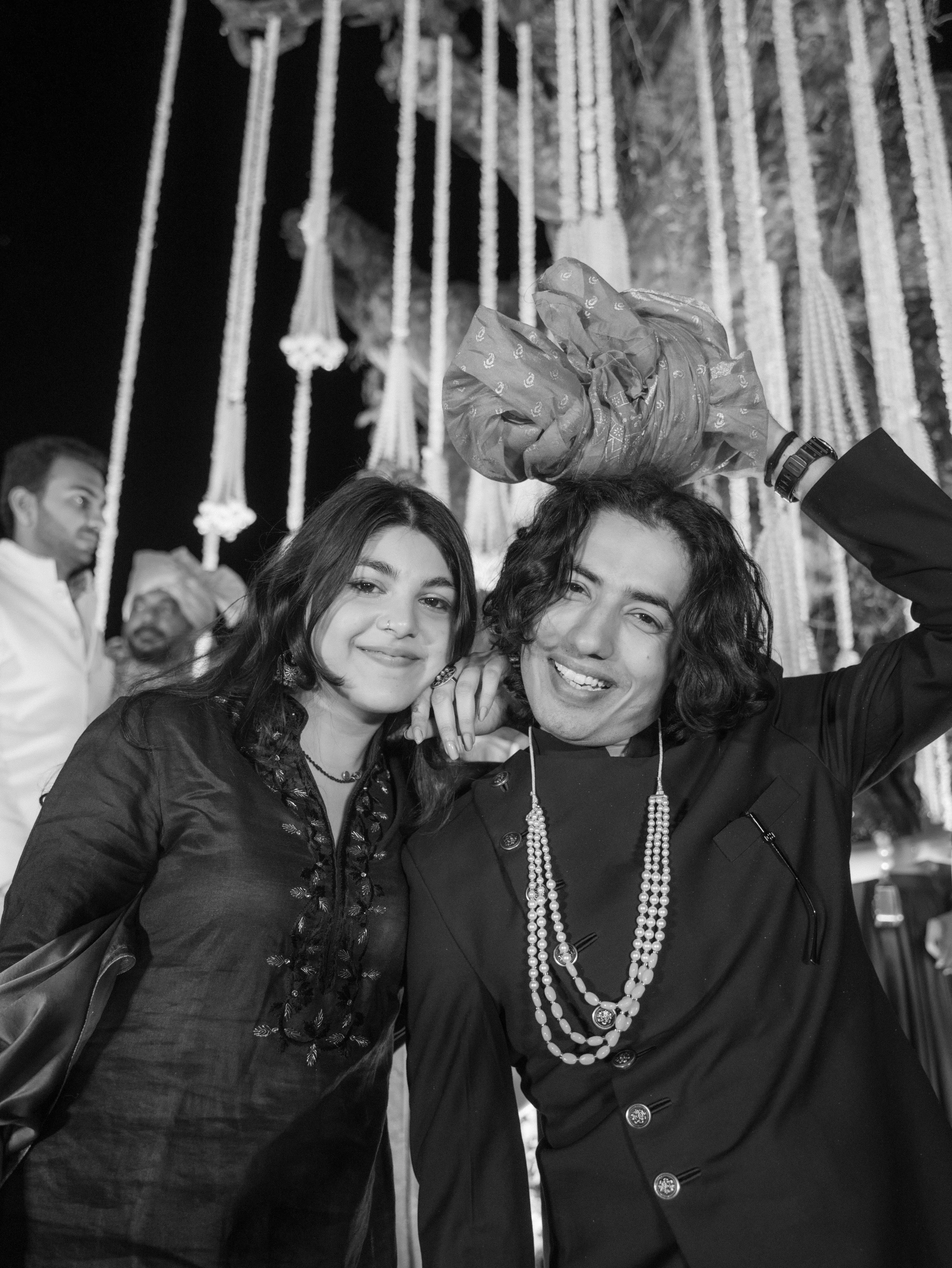 Two women smiling and taking a selfie, with one woman holding a wrapped gift. The background features hanging decorative elements, possibly at a celebration or event.