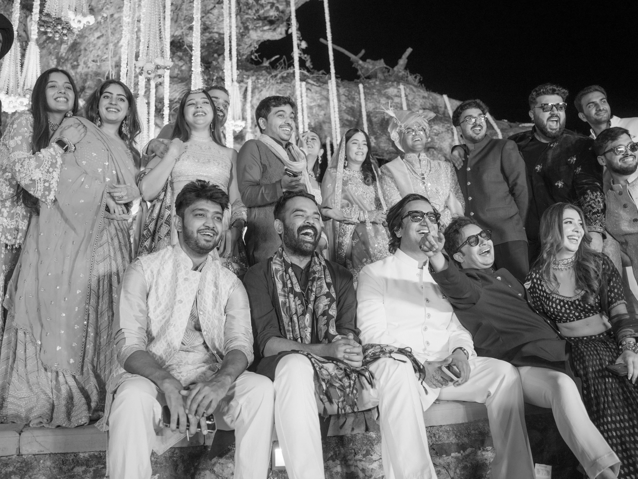 A group of people at night, dressed in traditional Indian clothing, sitting and smiling together, possibly at a celebration or wedding.