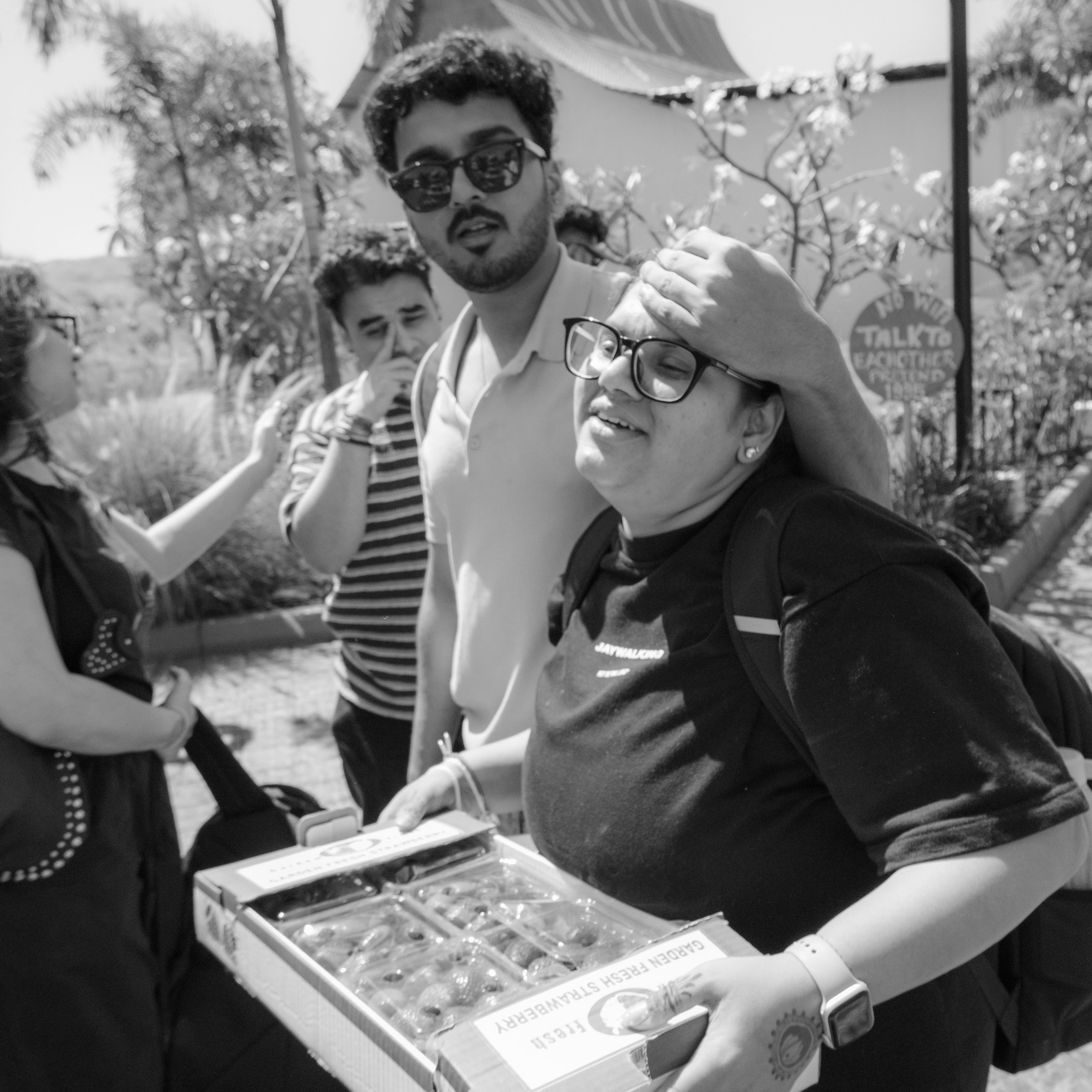 A group of people gathered outdoors around a table with a box of chocolates. One man with sunglasses and a beard has his arm around a woman wearing glasses. Another person in the background looks on. Trees and a sign are visible in the background.