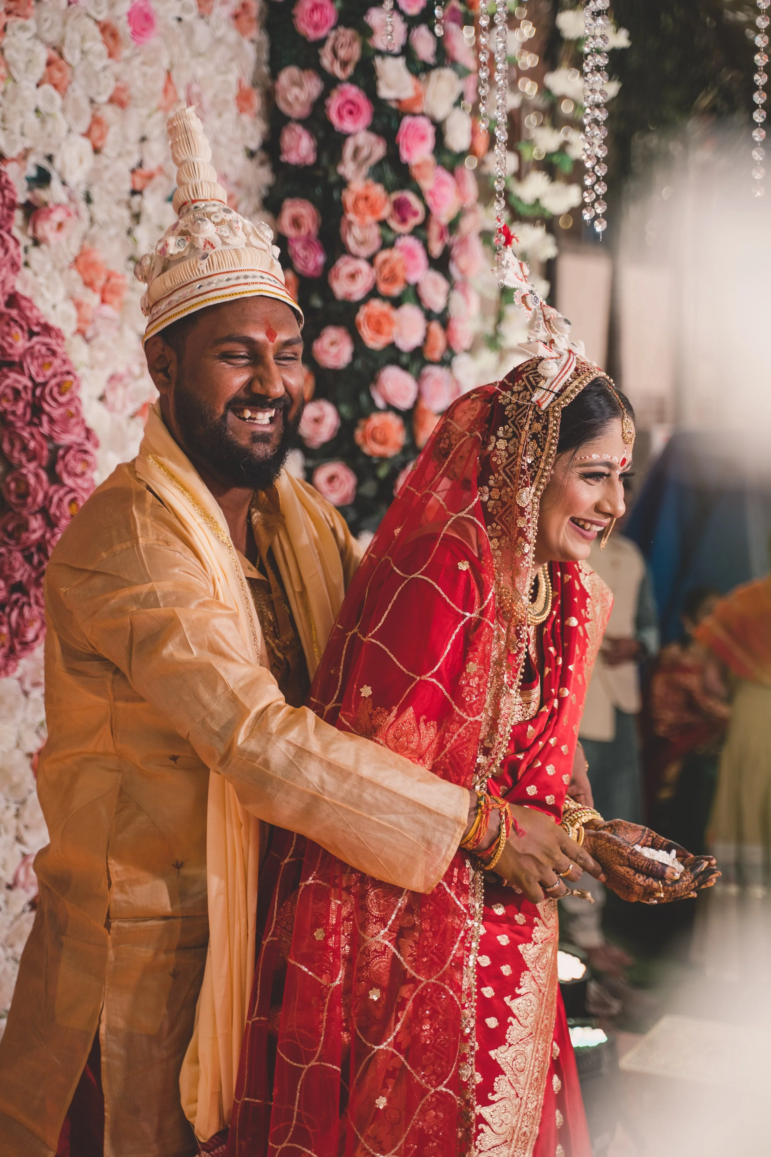A joyful groom and bride at their wedding, wearing traditional Indian attire, holding hands, with a floral backdrop of pink, white, and peach roses and hanging crystal decorations, smiling and laughing.
