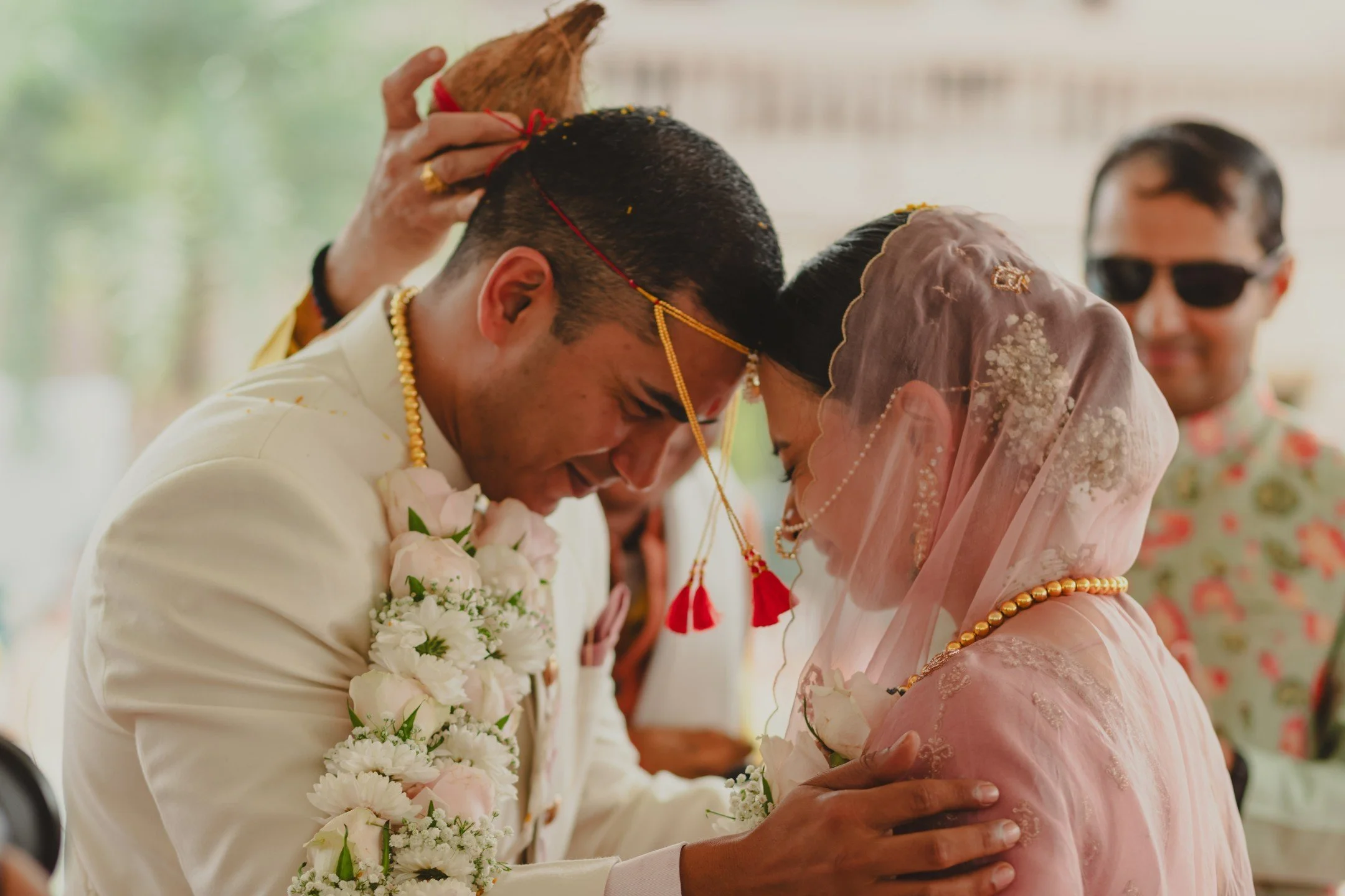 A couple dressed in traditional Indian wedding attire bow their heads together during a ceremony, with a man in the background wearing sunglasses smiling.