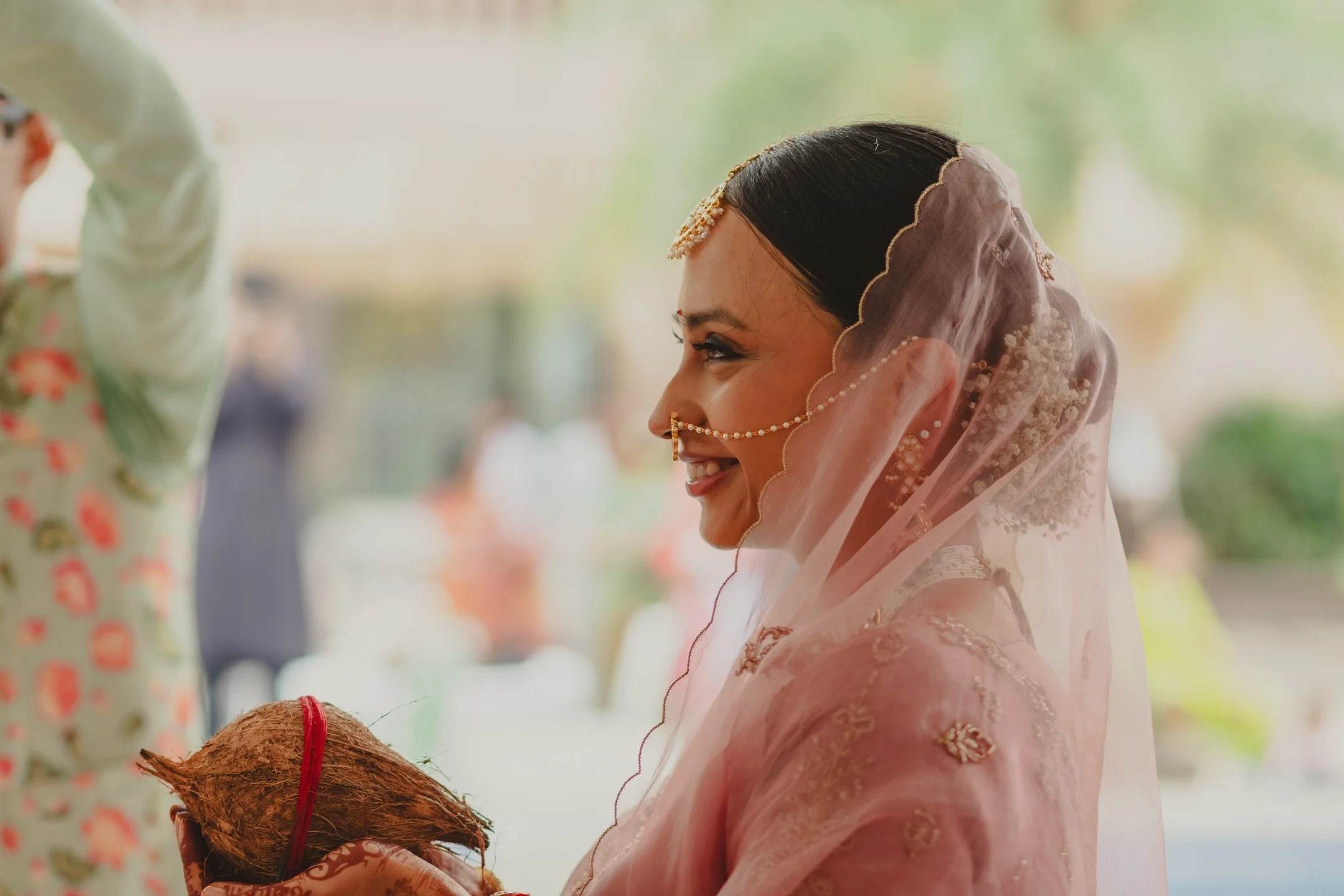 A woman dressed in traditional pink Indian attire and jewelry, smiling while holding a coconut during a cultural or religious ceremony.