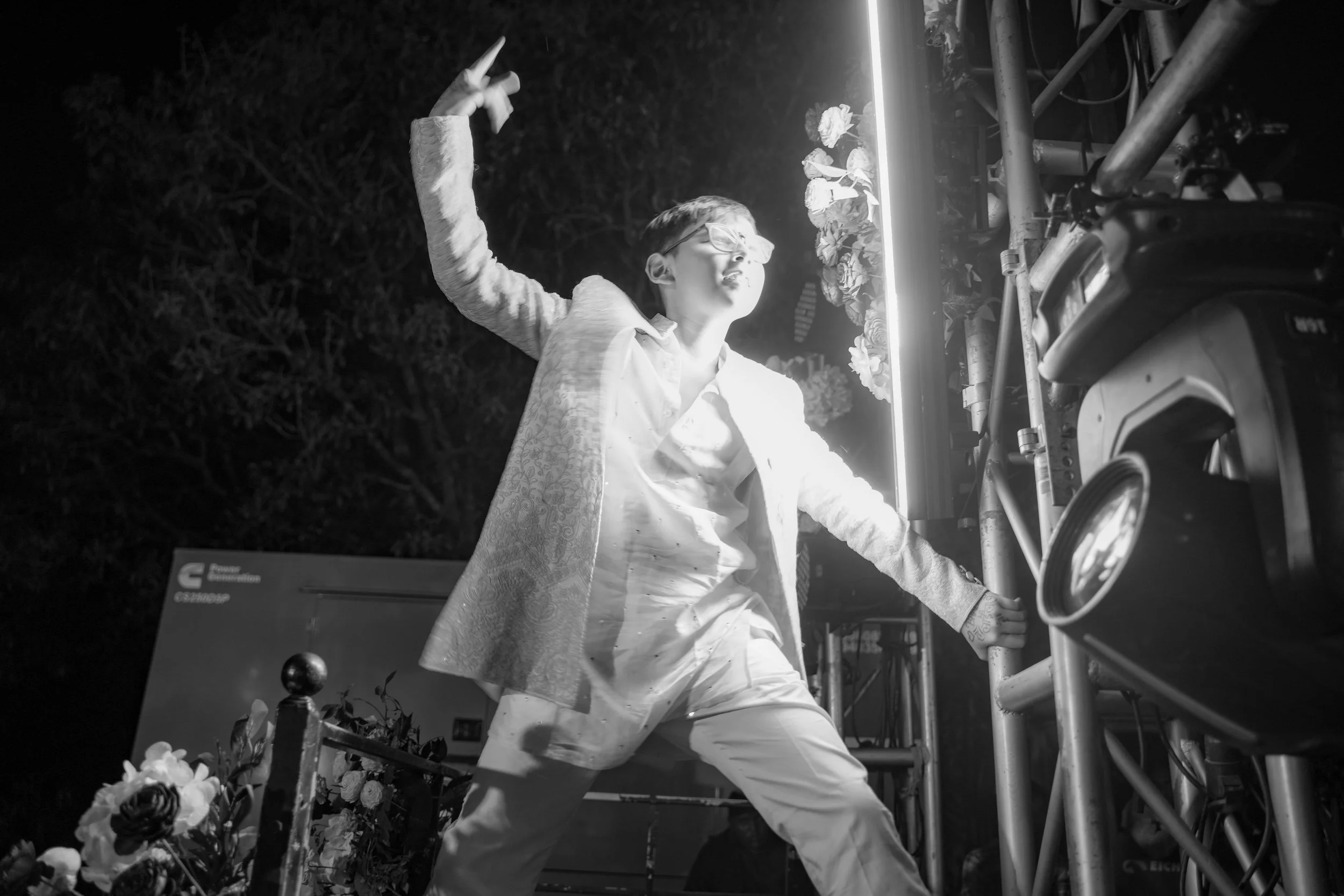 Person with short hair and glasses, wearing a patterned shirt and white pants, standing outdoors at night, reaching up with their right arm, near a flower arrangement and a structural frame with bright lighting.