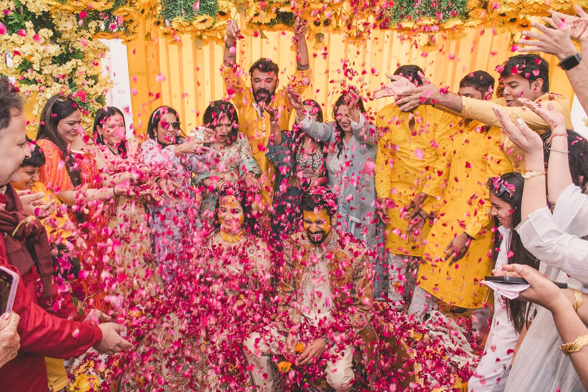 A group of people celebrating a wedding, throwing pink rose petals and smiling. The background is decorated with yellow flowers and yellow drapes. The bride and groom are in the center, surrounded by friends and family.