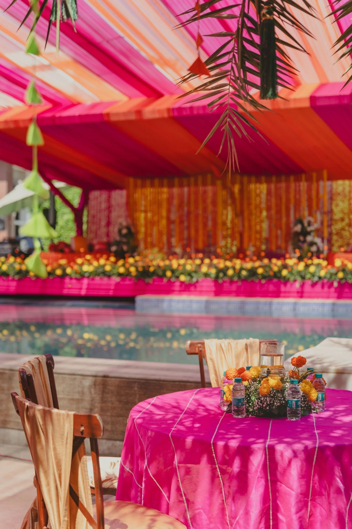 Decorated outdoor setting with pink tablecloth, floral centerpiece, water bottles, and chairs. In the background, colorful drapes and hanging decorations create a festive atmosphere.