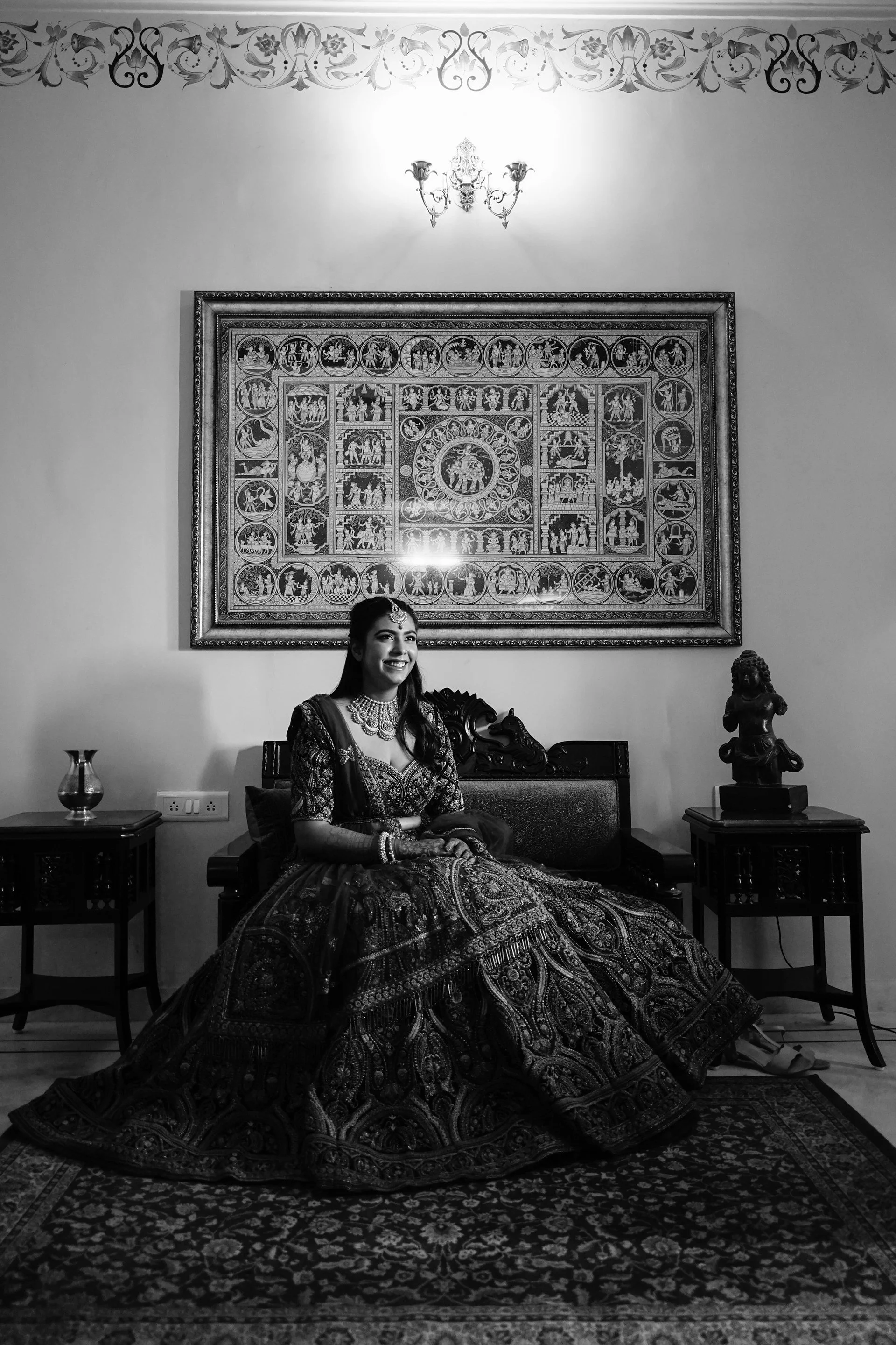 A woman in traditional Indian attire sitting on a sofa in a richly decorated room with a large framed artwork on the wall behind her, flanked by two side tables with decorative items, and a patterned rug on the floor.
