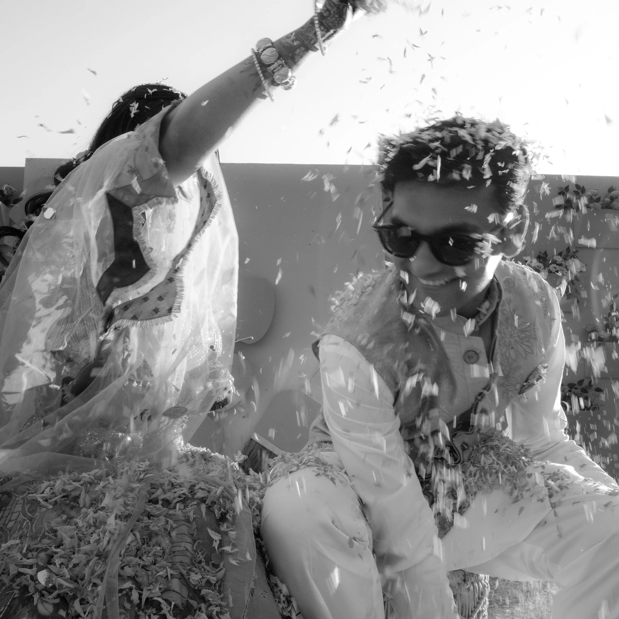A black and white photograph of a bride with an updo hairdo, wearing glasses, and a wedding gown, throwing confetti or rice. The scene appears to be a wedding celebration outdoors.