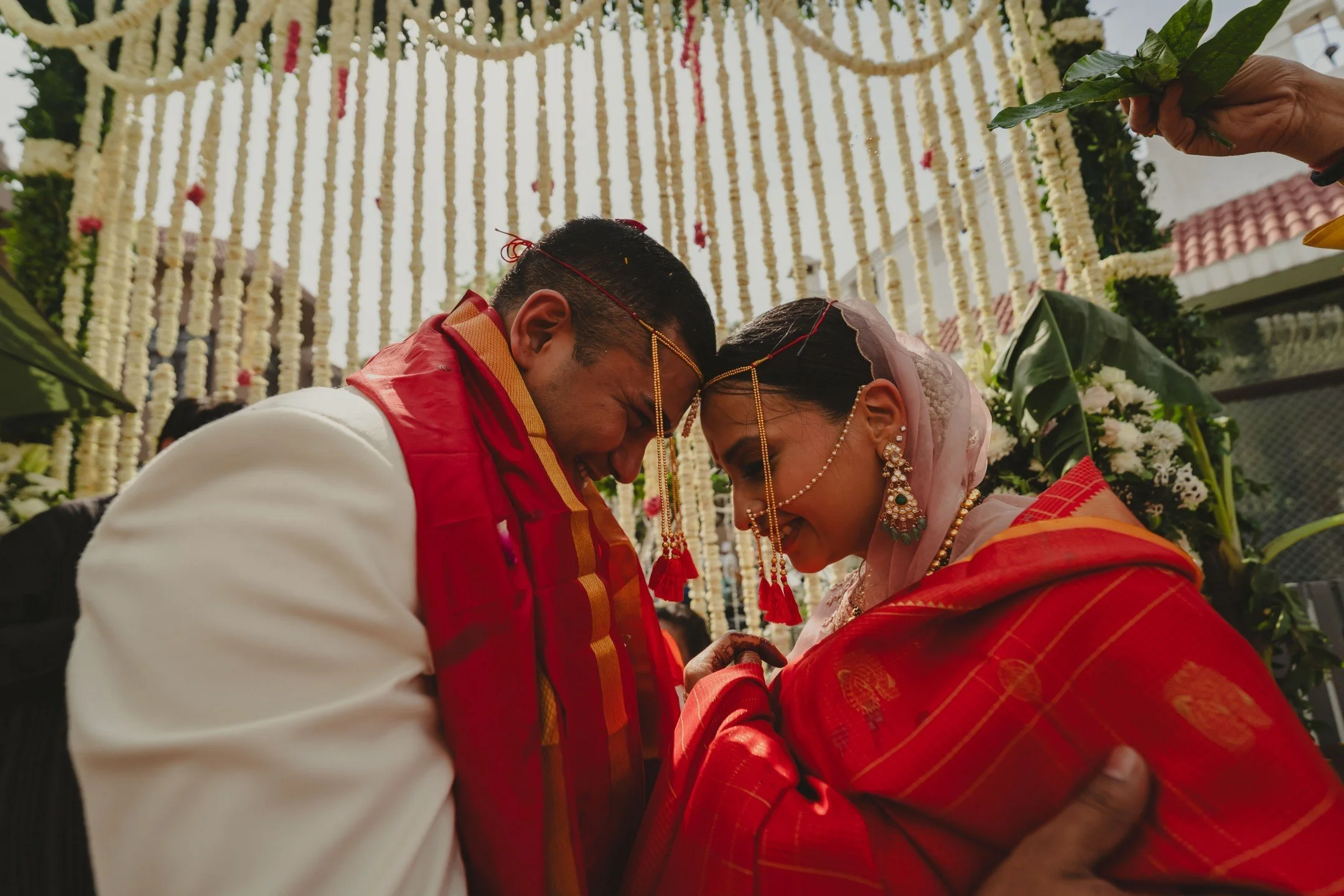 A Hindu wedding ceremony with a couple bowing with foreheads touching, adorned in traditional red and gold attire, with flower decorations overhead.