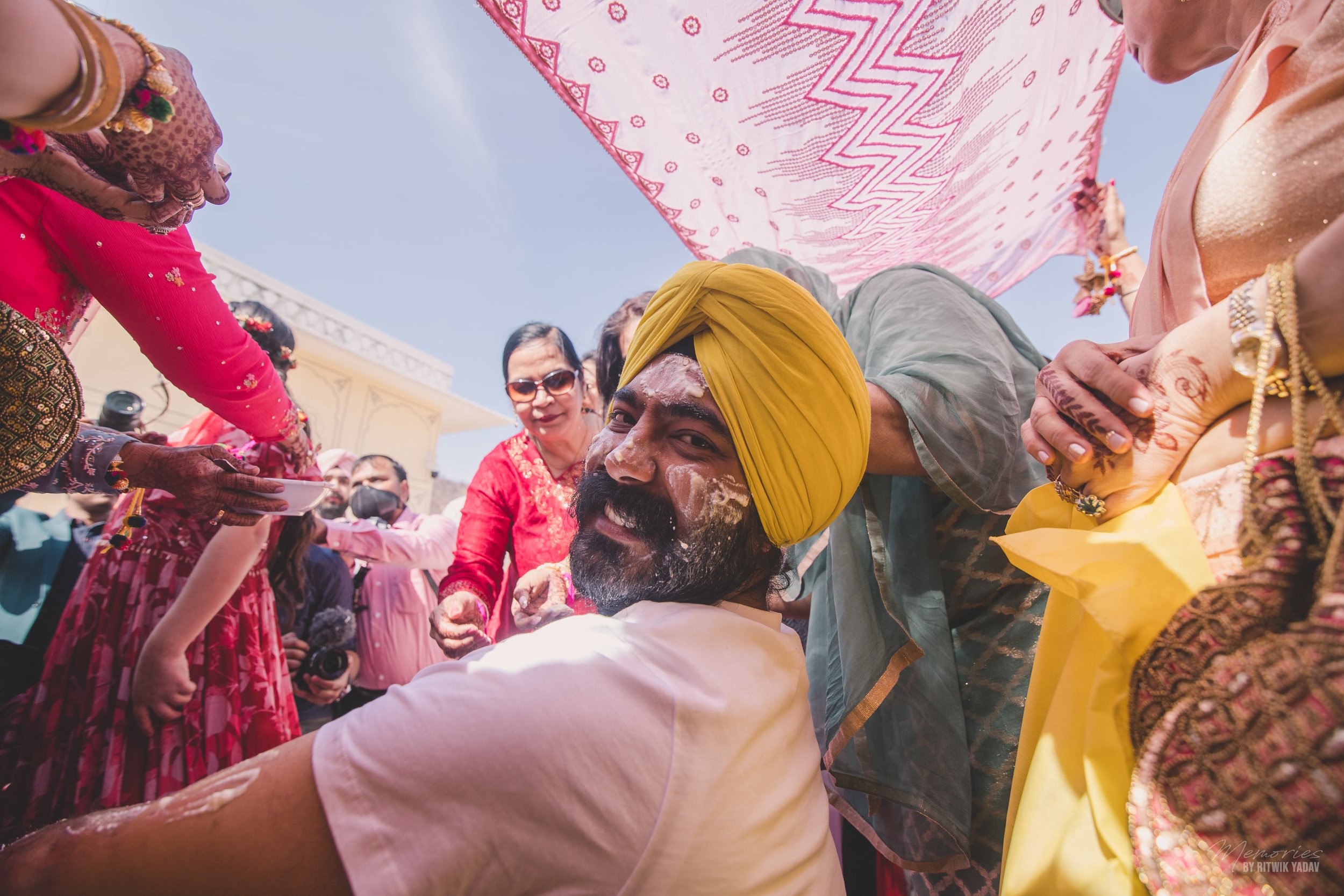 A group of people gathered outdoors, with a man wearing a yellow turban and traditional attire. He is smiling and leaning forward, engaging with the crowd. Women around him are dressed in colorful sarees and traditional clothing, some with intricate jewelry. The sky is clear and sunny, indicating a festive or cultural event.