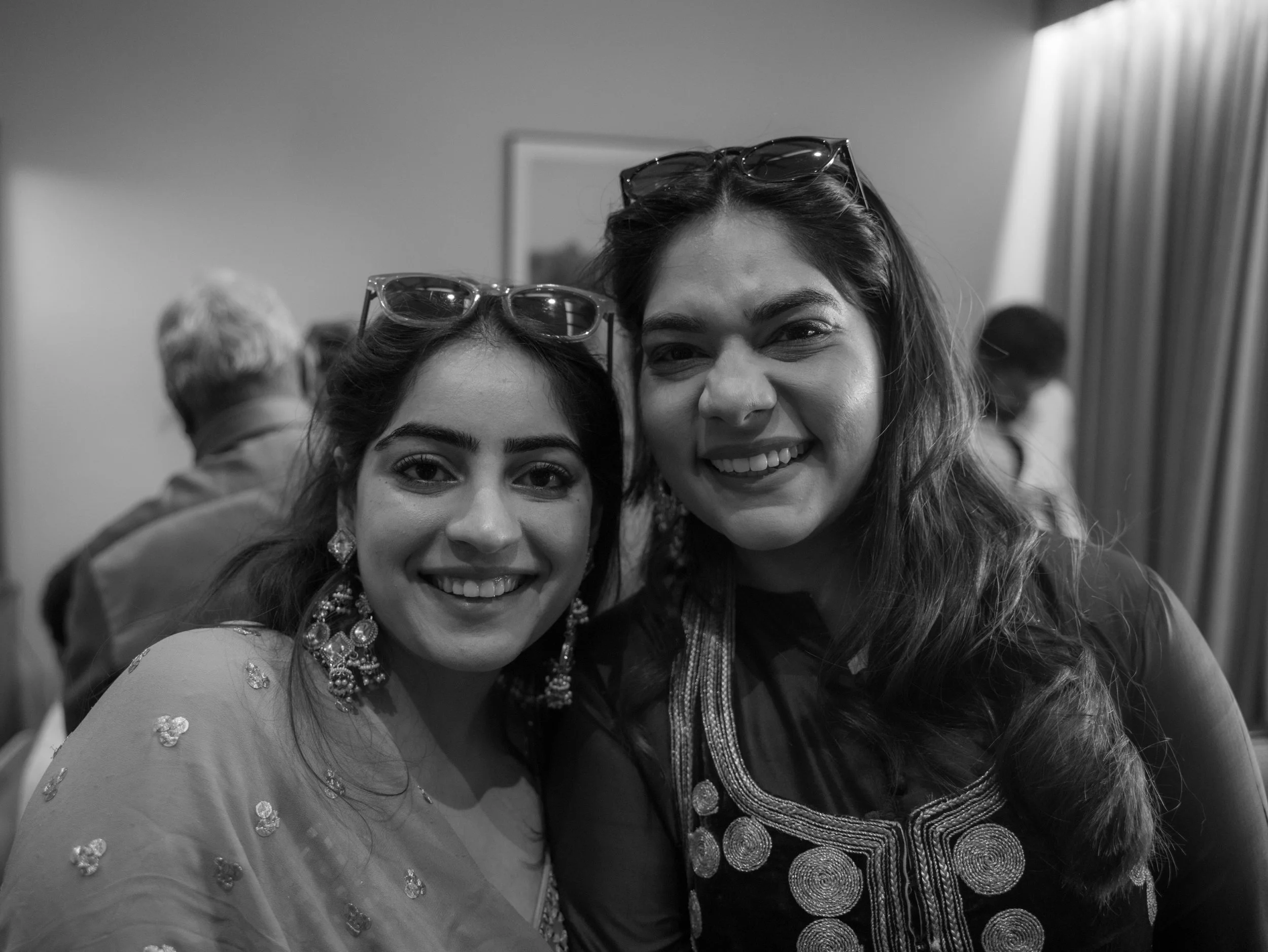 Two women smiling and wearing glasses on their heads, close-up selfie at an indoor event.
