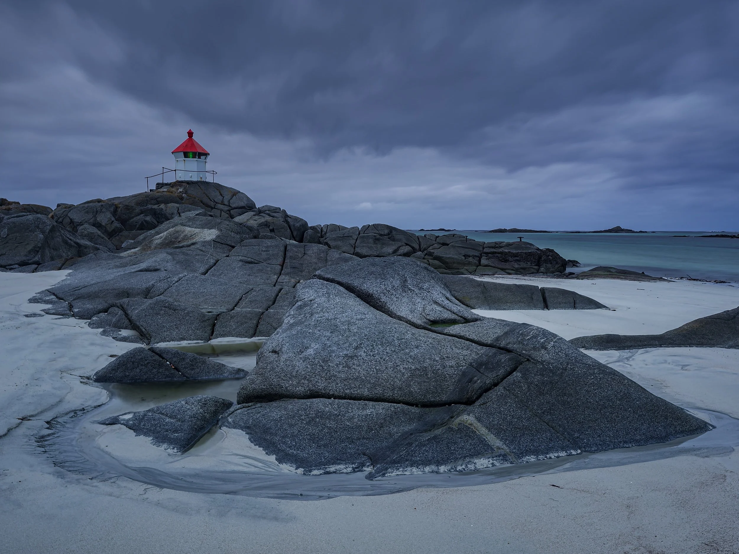 A rocky beach scene with large gray rocks in the foreground, a small lighthouse with a red roof perched on the rocks, and dark clouds overhead with a view of the ocean and distant landmasses in the background.