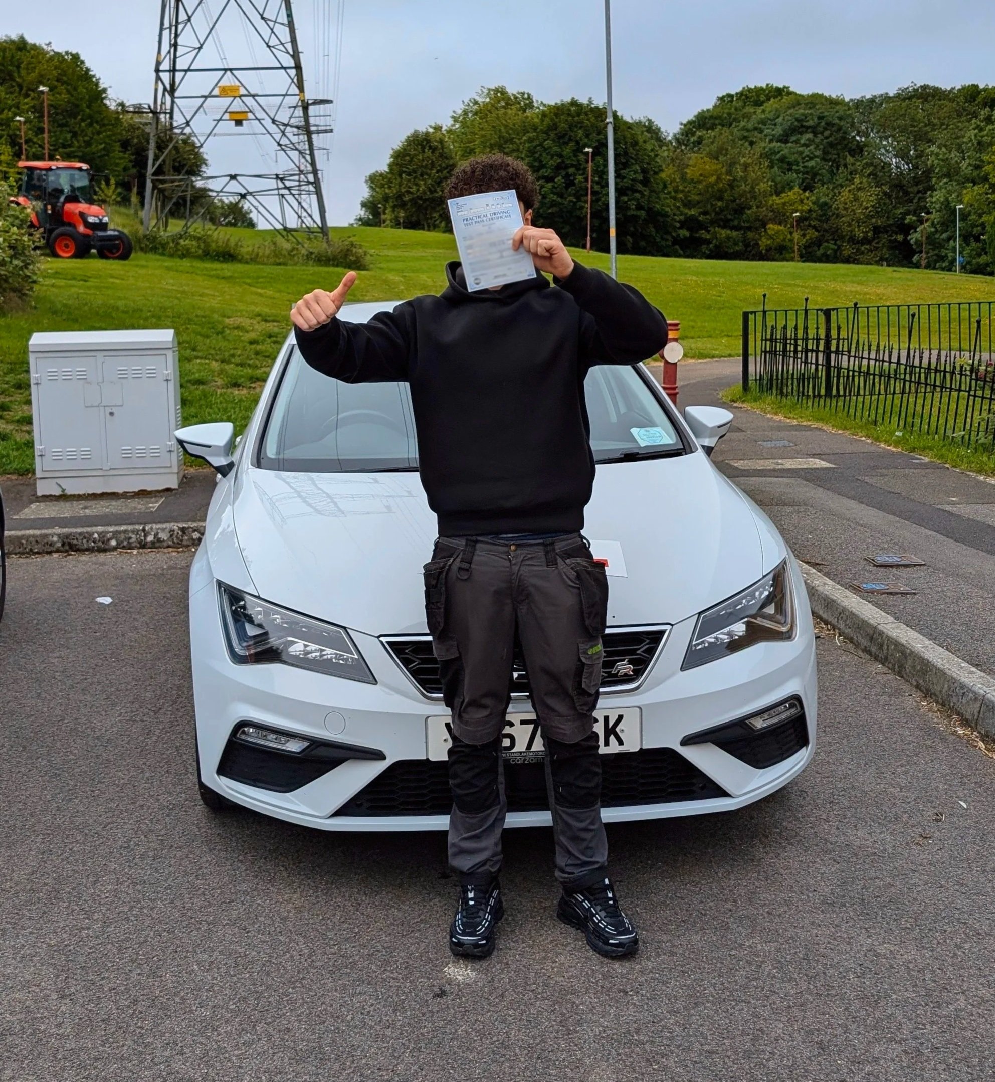 Person standing in front of a white car, holding a driver's license covering their face, giving a thumbs-up with their left hand, outdoors in a parking lot with green trees, utility pole, and a tractor in the background.