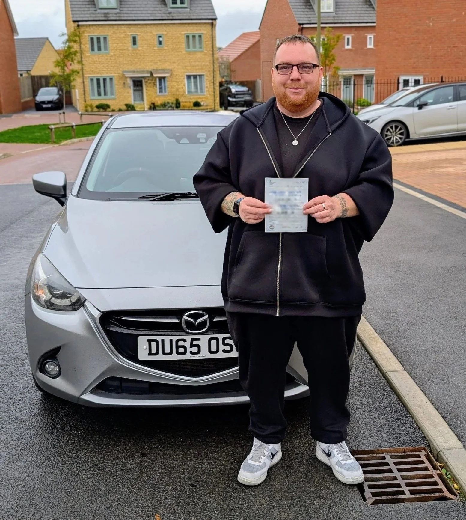A man with glasses and a beard holding a license standing in front of a silver Mazda car with a blurred license plate that reads 'DU65 OSP'. He is wearing a black jacket, black pants, white sneakers, a necklace, and has tattoos on his left arm. The background features residential houses, parked cars, and a street in a suburban neighborhood on a cloudy day.