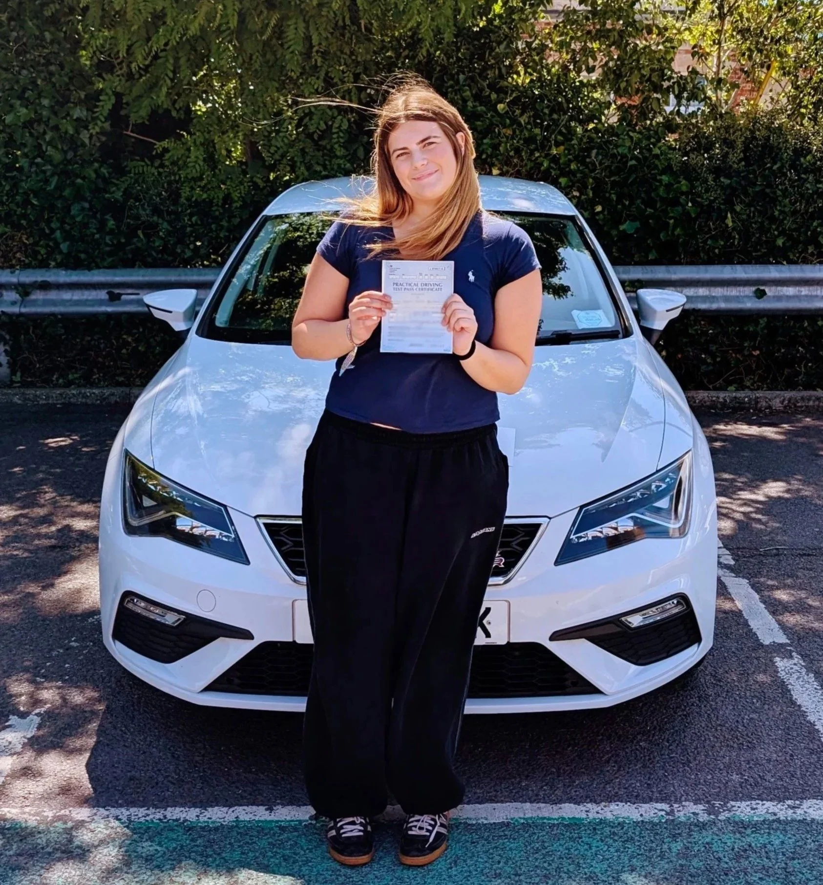 Young woman standing in front of a white car, holding a practical driving test pass certificate, outdoors on a sunny day.