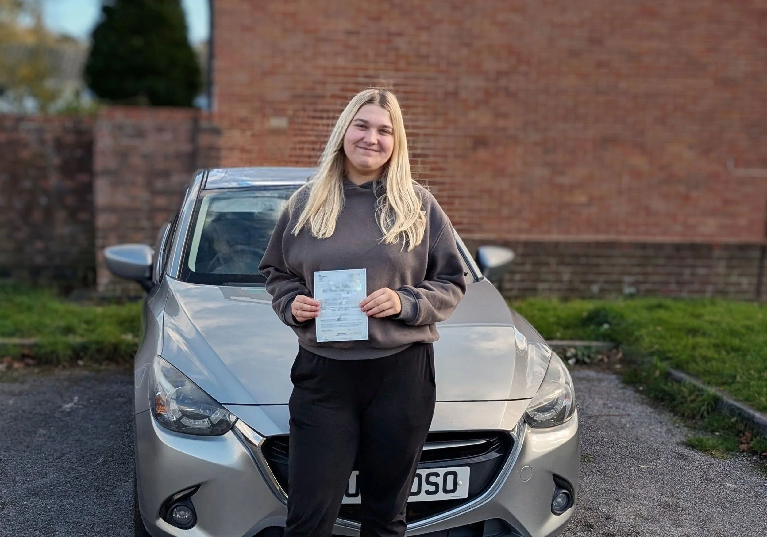 A young woman with long blond hair, smiling, holding a document while standing in front of a silver car parked outdoors, with a brick wall and some greenery in the background.