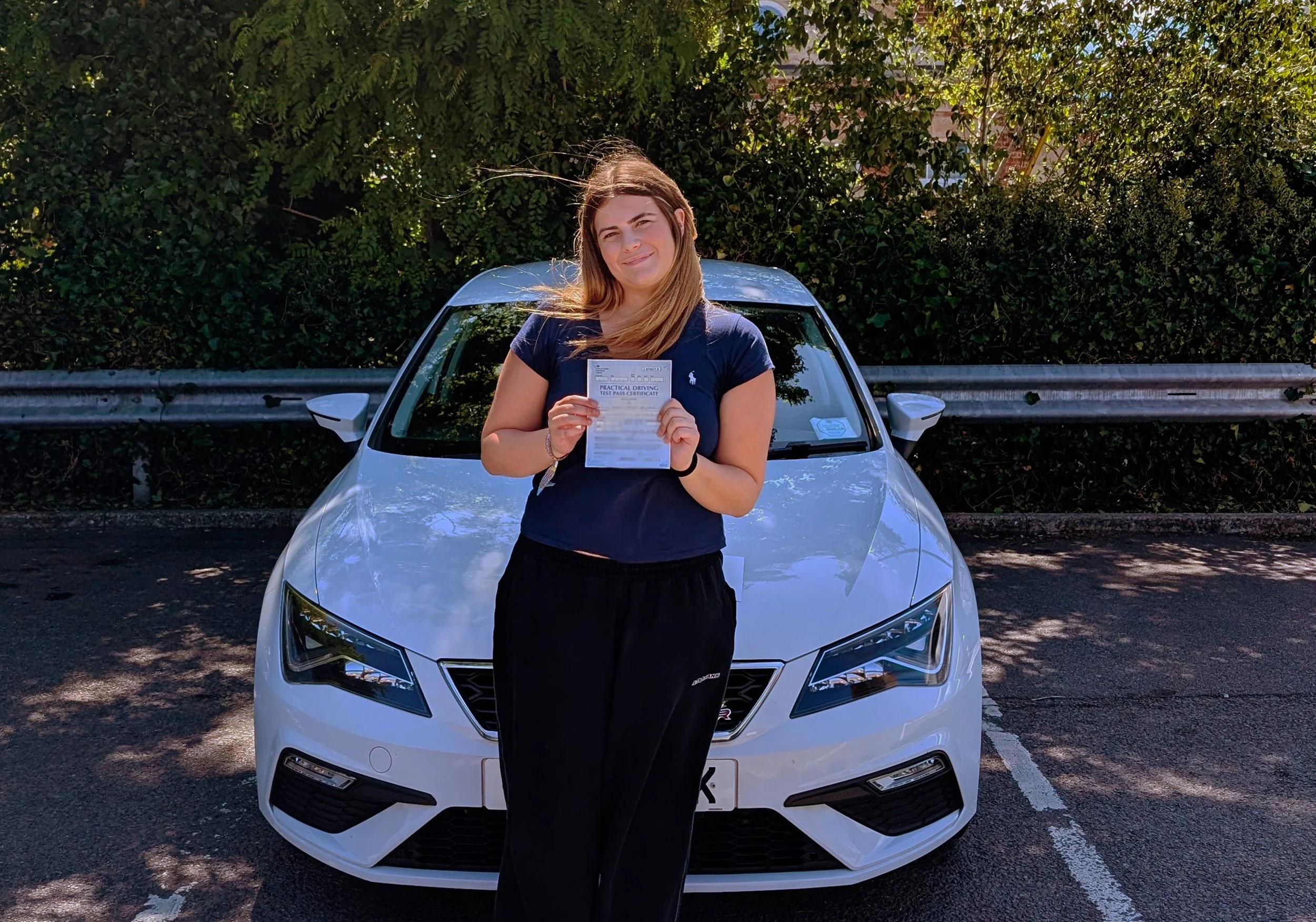 A woman standing in front of a white car, holding a practical driving test pass certificate, outdoors near a green hedge.