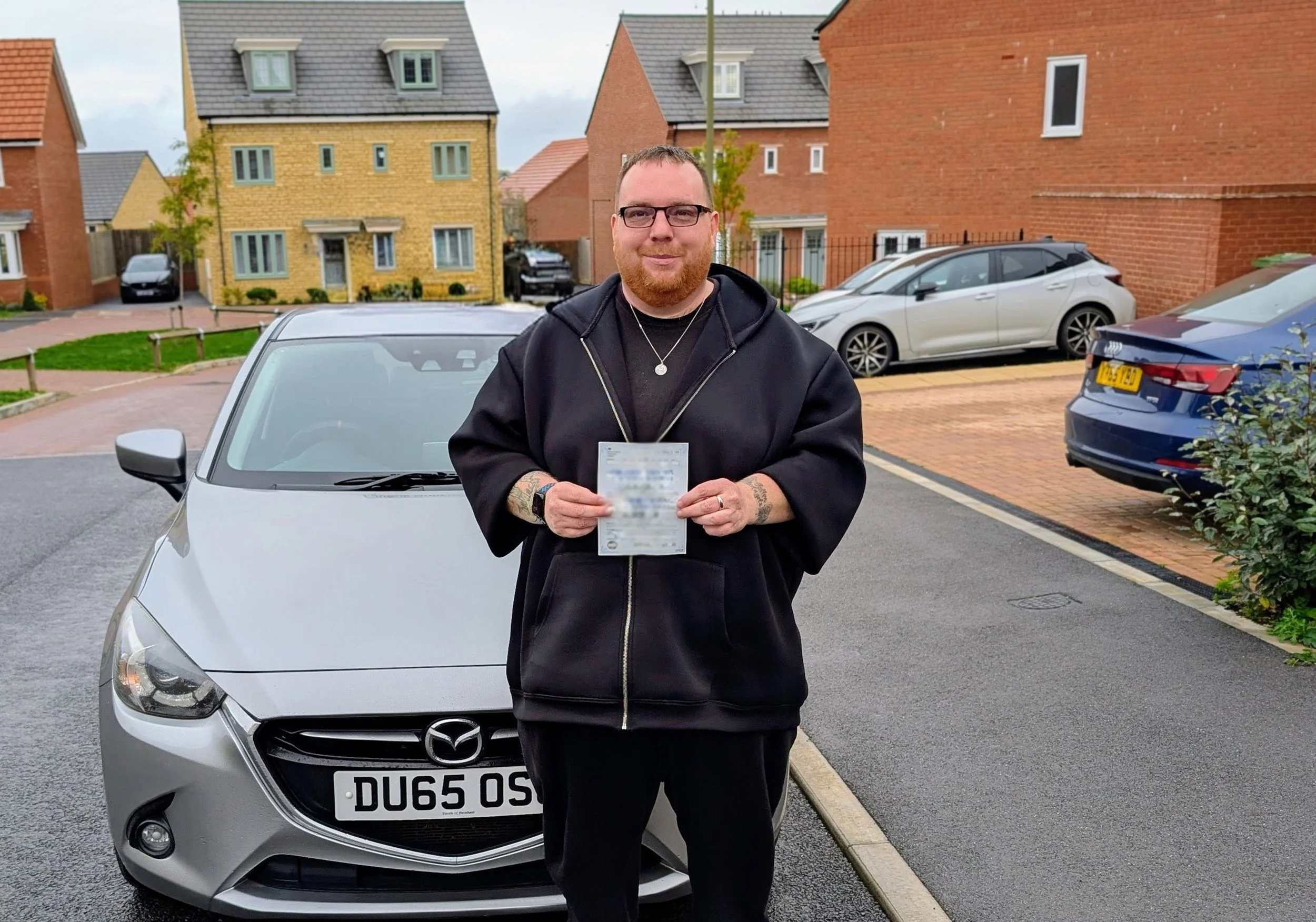 A man with glasses and a beard standing in front of a silver Mazda car in a residential neighborhood, holding a document, with houses and parked cars visible in the background.
