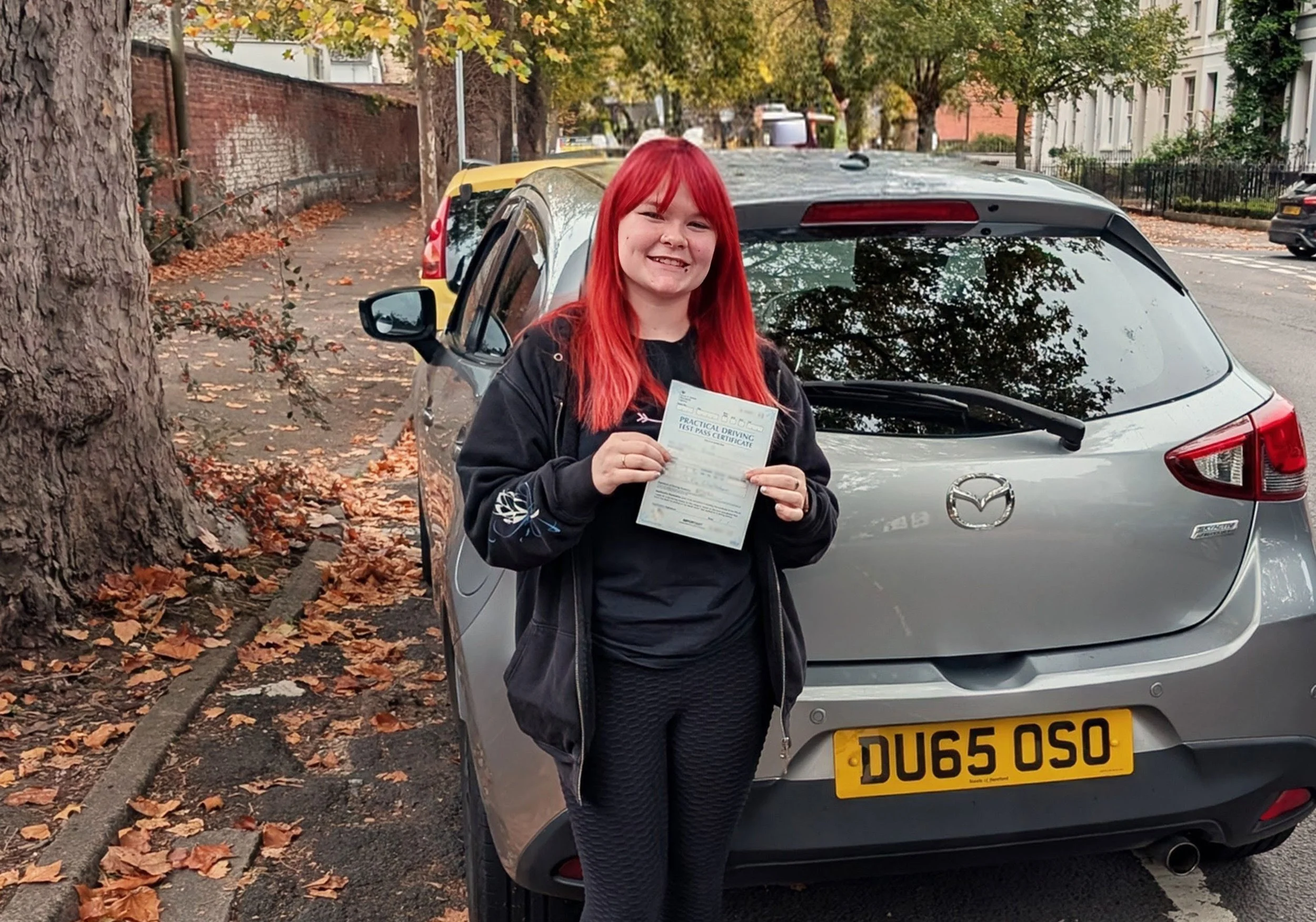 A young woman with bright red hair smiling and holding a practical driving test pass certificate, standing in front of a silver Mazda car parked on a street with autumn leaves on the ground.