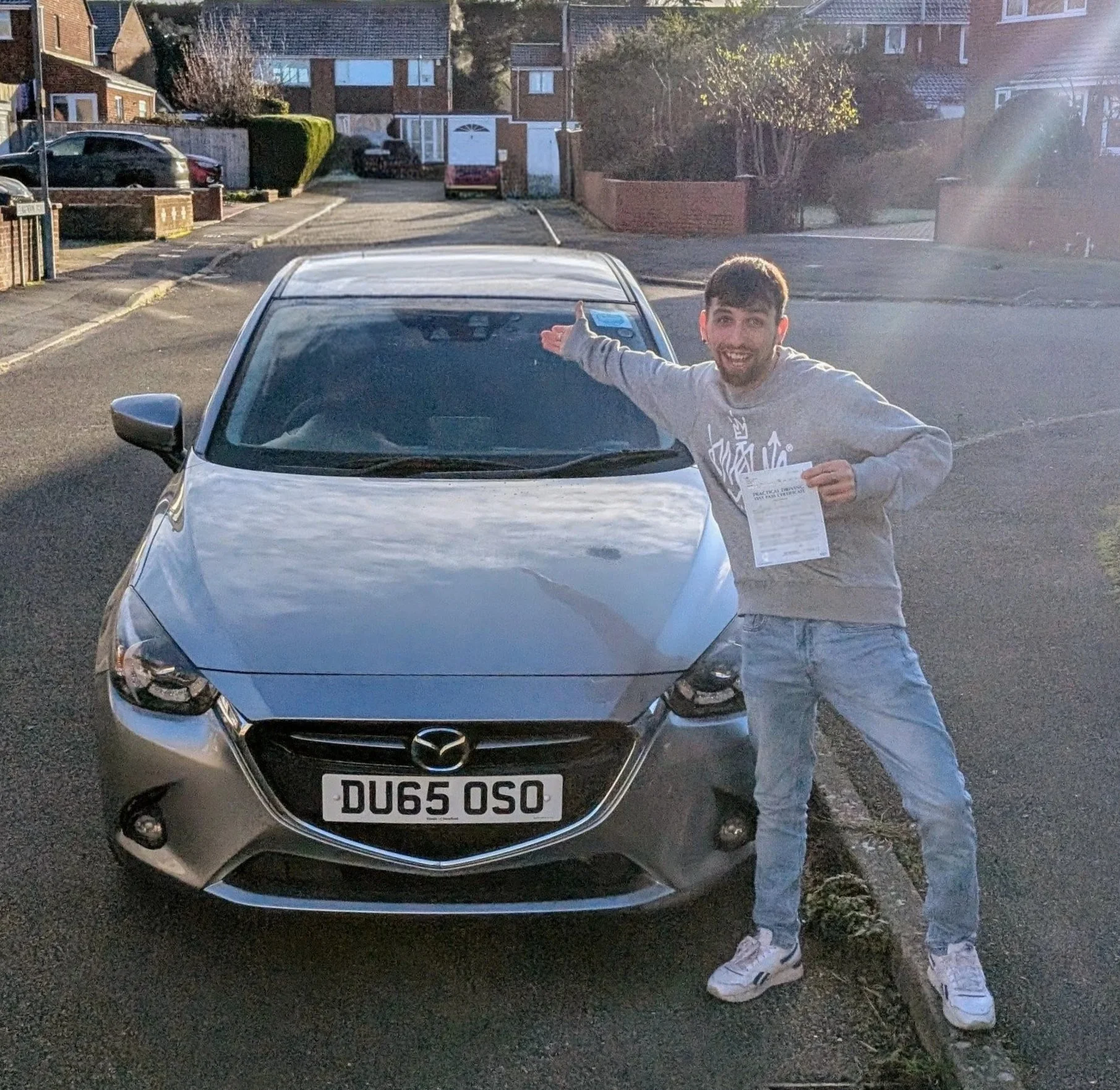 A young man with short dark hair and a beard, wearing a gray hoodie and light-colored jeans, standing next to a silver Mazda car. He is smiling, holding a piece of paper in one hand and gesturing towards the car with his other arm extended. The car's license plate reads 'DU65 OSO'. The scene is outdoors in a residential area with houses, trees, and parked cars visible in the background, illuminated by natural sunlight.
