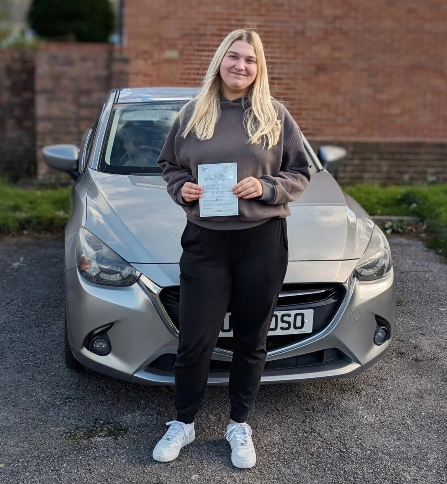 A young woman with long blonde hair, wearing a gray hoodie and black pants, stands in front of a silver car holding a piece of paper, smiling.