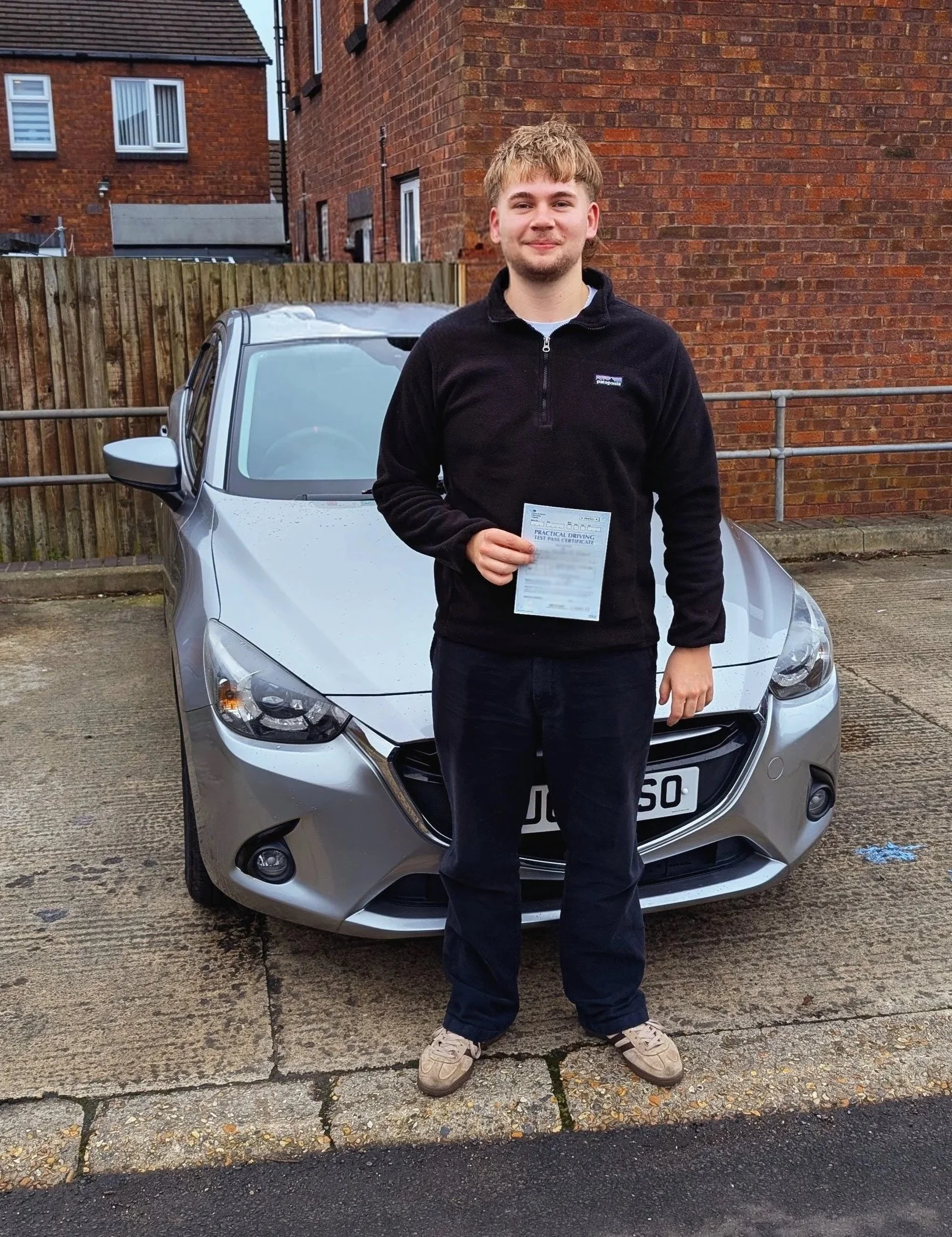 A young man standing in front of a silver car on a paved surface, holding a driving test certificate, with a red brick building and a wooden fence in the background.