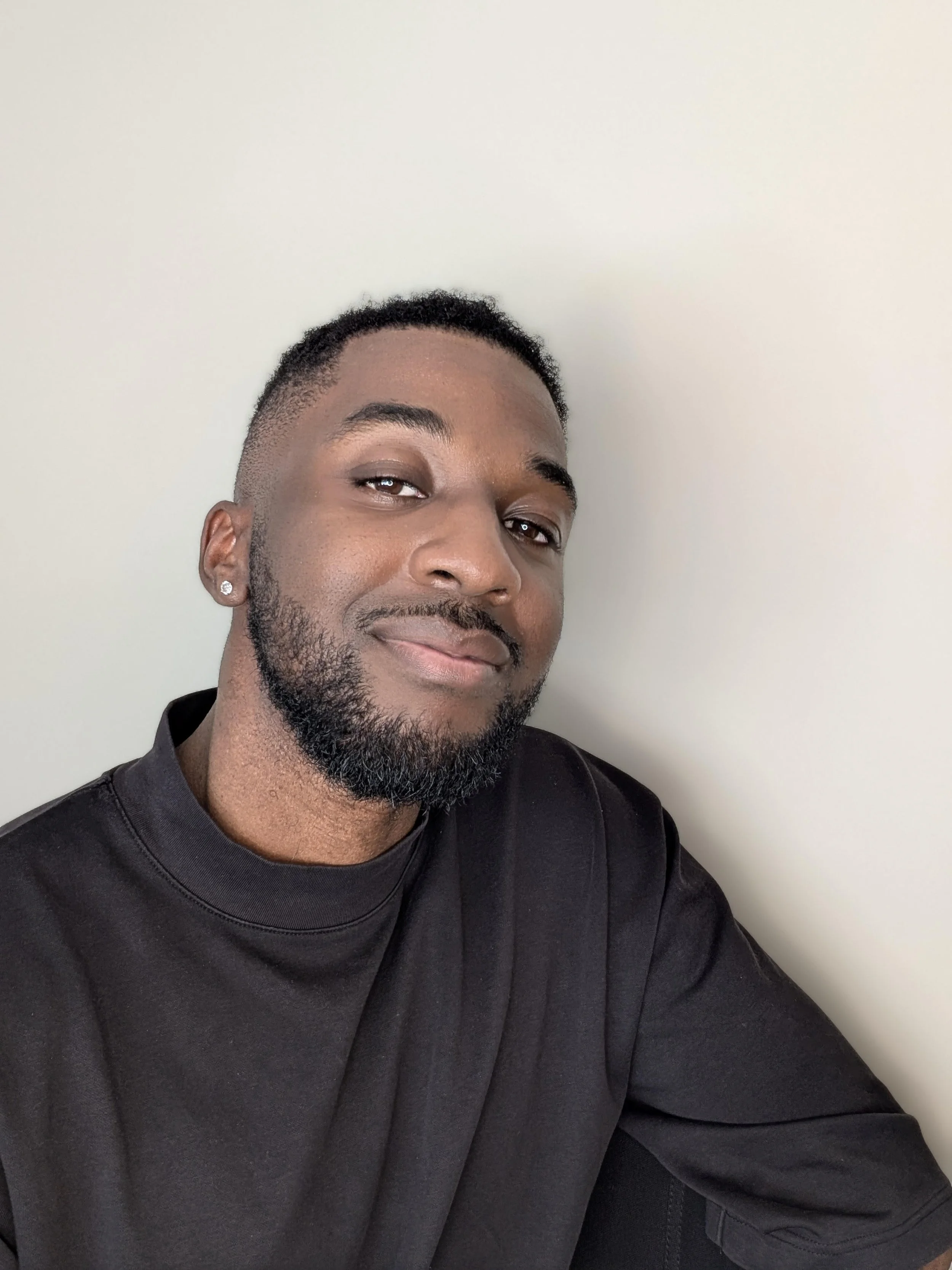 Close-up of a young man with short curly hair, a neatly trimmed beard, wearing earrings and a black sweatshirt, smiling slightly, against a plain light-colored wall.