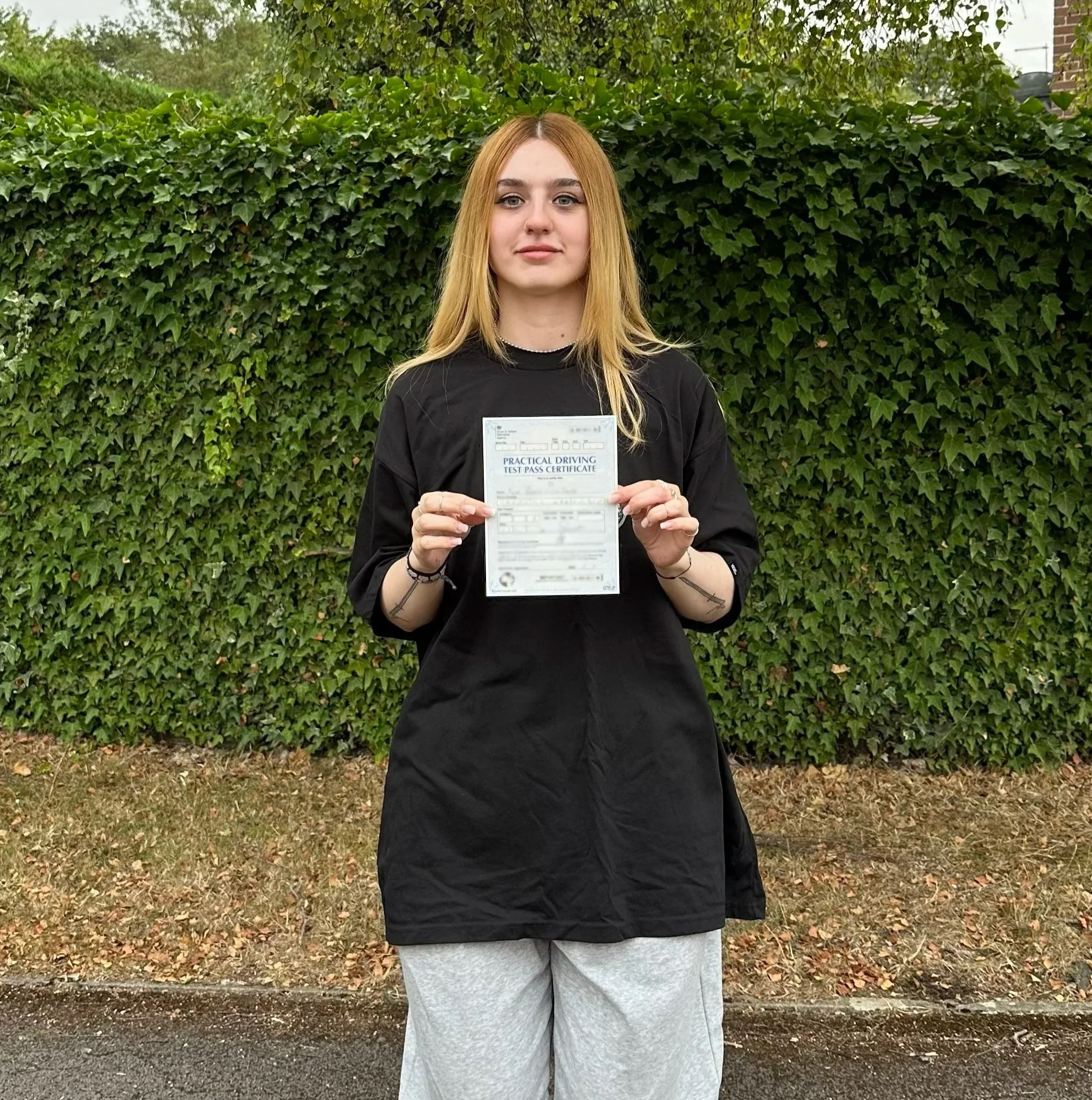 A young woman with long blonde hair holding a practical driving test pass certificate standing outdoors in front of a green leafy hedge.