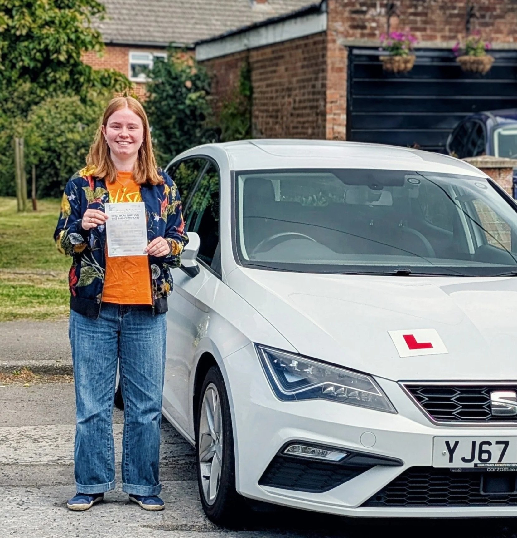A young woman with red hair, wearing a floral jacket and jeans, holding a piece of paper, standing next to a white electric car with a learner's 'L' sign on the front, in a residential area with brick houses and greenery.